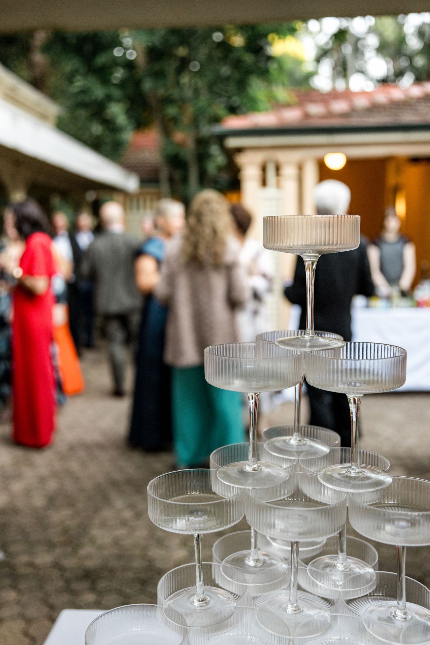 Champagne tower with coupe glasses at elegant outdoor wedding reception with guests mingling behind.