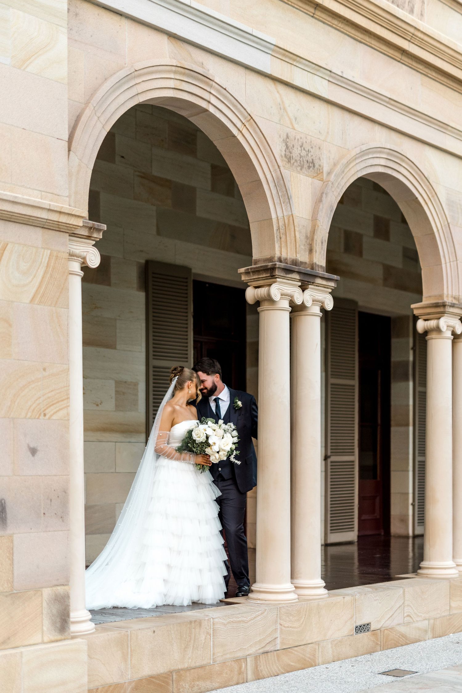 Bride in layered tulle gown and groom posing under sandstone arches at historic venue.