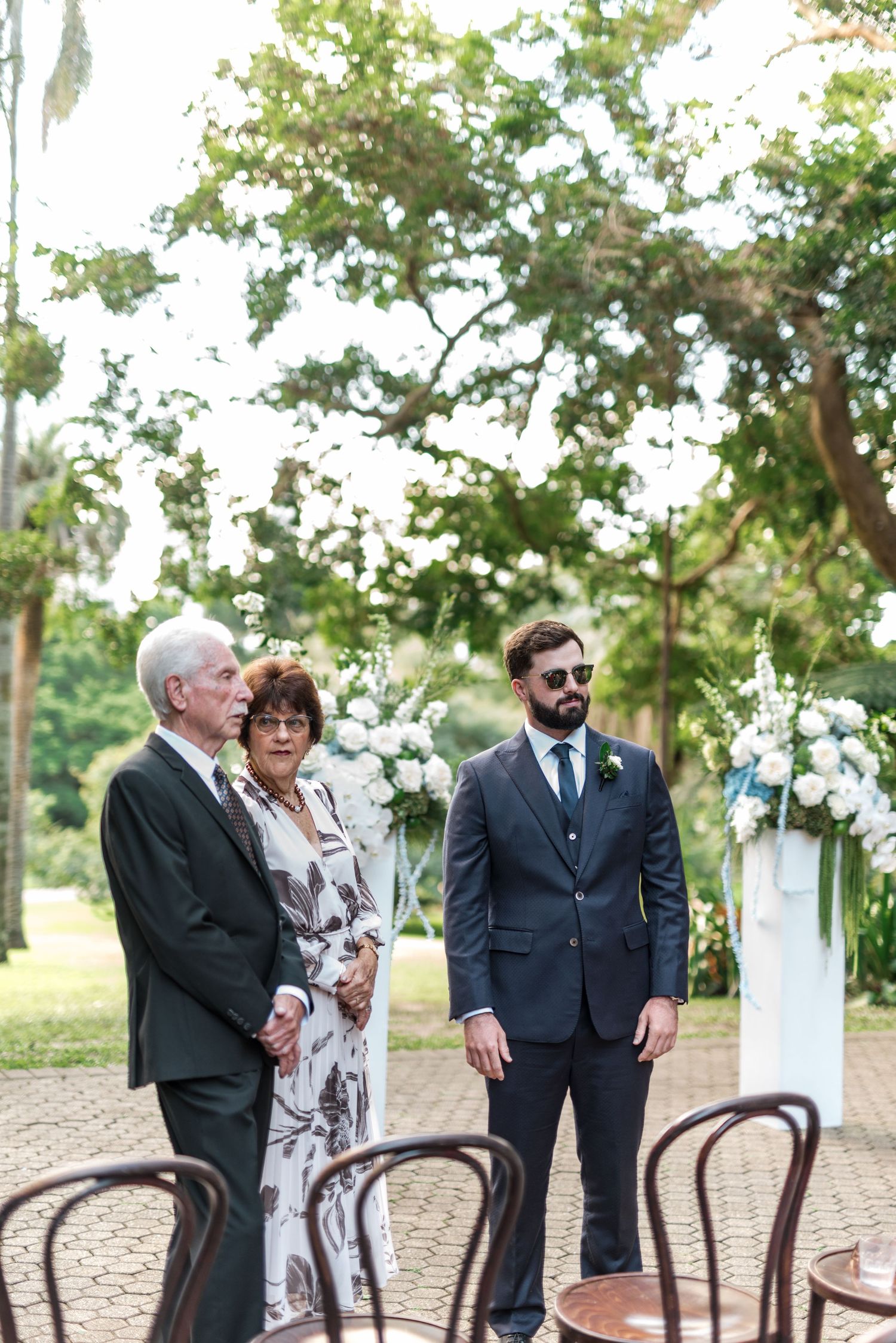 Groom in navy suit stands with elderly couple at outdoor wedding ceremony altar.