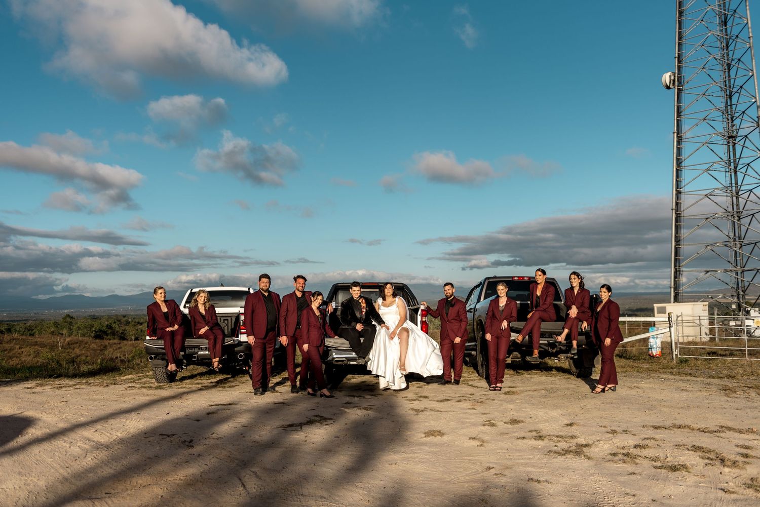 Wedding party in burgundy suits posing with bride and groom beside black trucks outdoors.