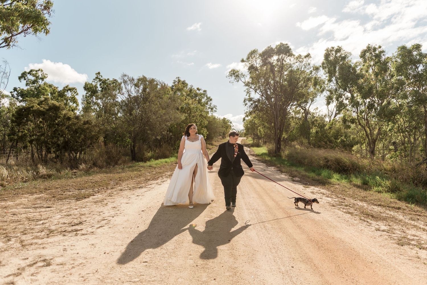 Bride and groom walk joyfully with their small black dog on a sunny rural dirt road.