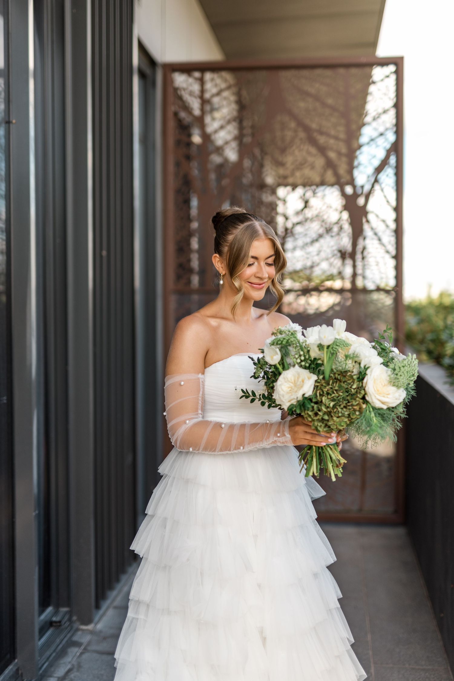 Bride in tiered tulle gown with sheer pearl sleeves holds white and green bouquet on sunlit balcony.