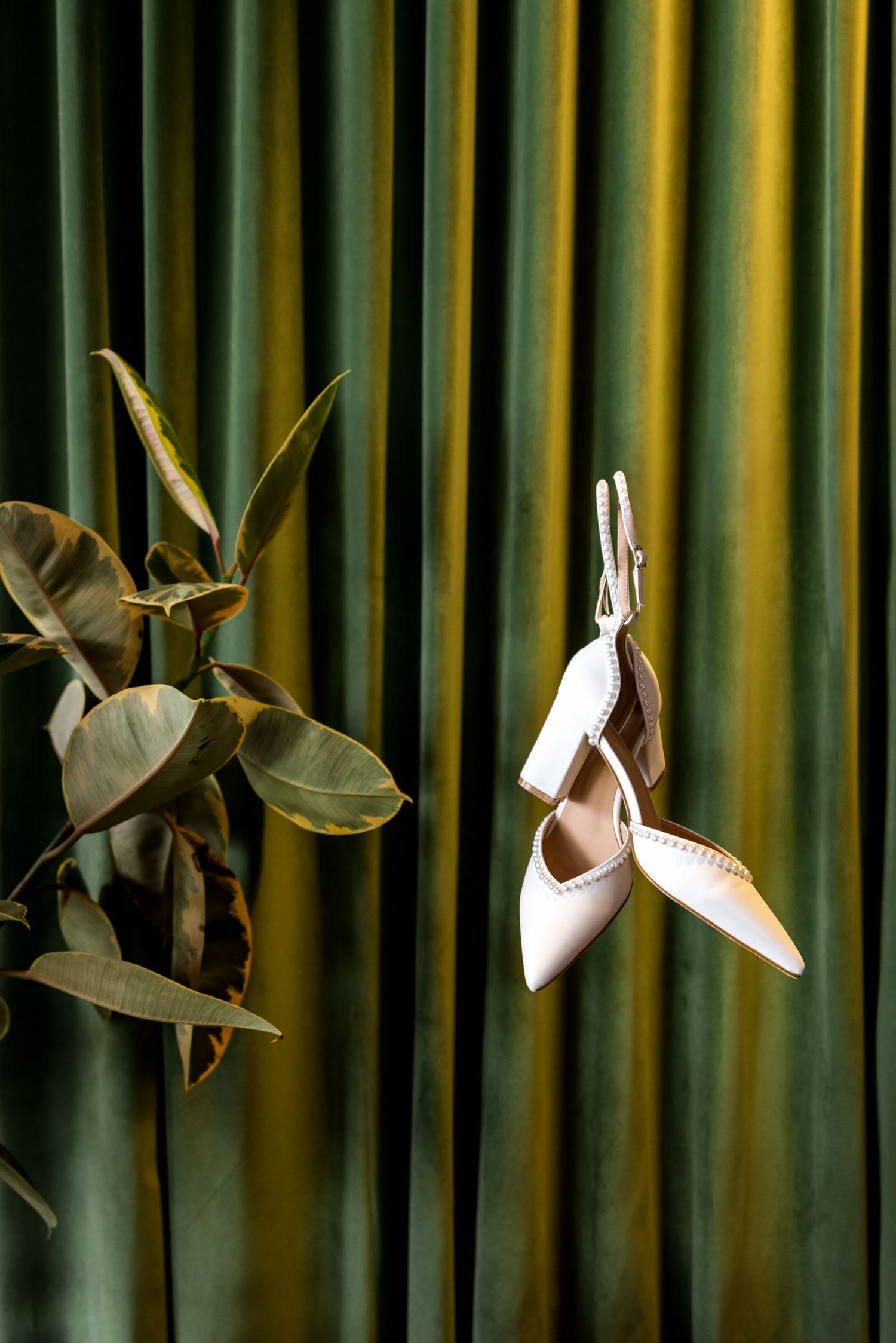 Elegant white pointed-toe bridal heels hanging against a green velvet curtain with tropical leaves.