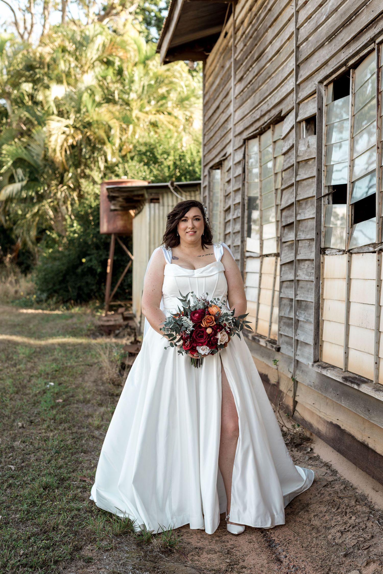 Bride in white satin gown with thigh slit holds a boho bouquet beside a rustic weathered building.