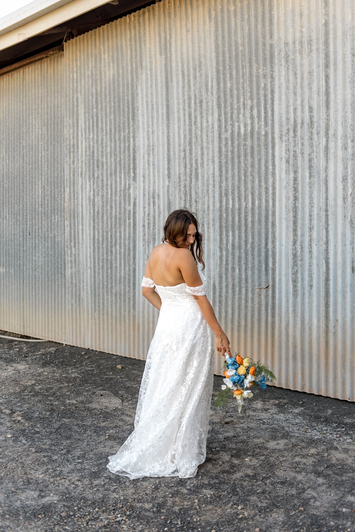 Bride in off-shoulder lace wedding gown holds colorful bouquet, posing with back turned near rustic corrugated metal wall.