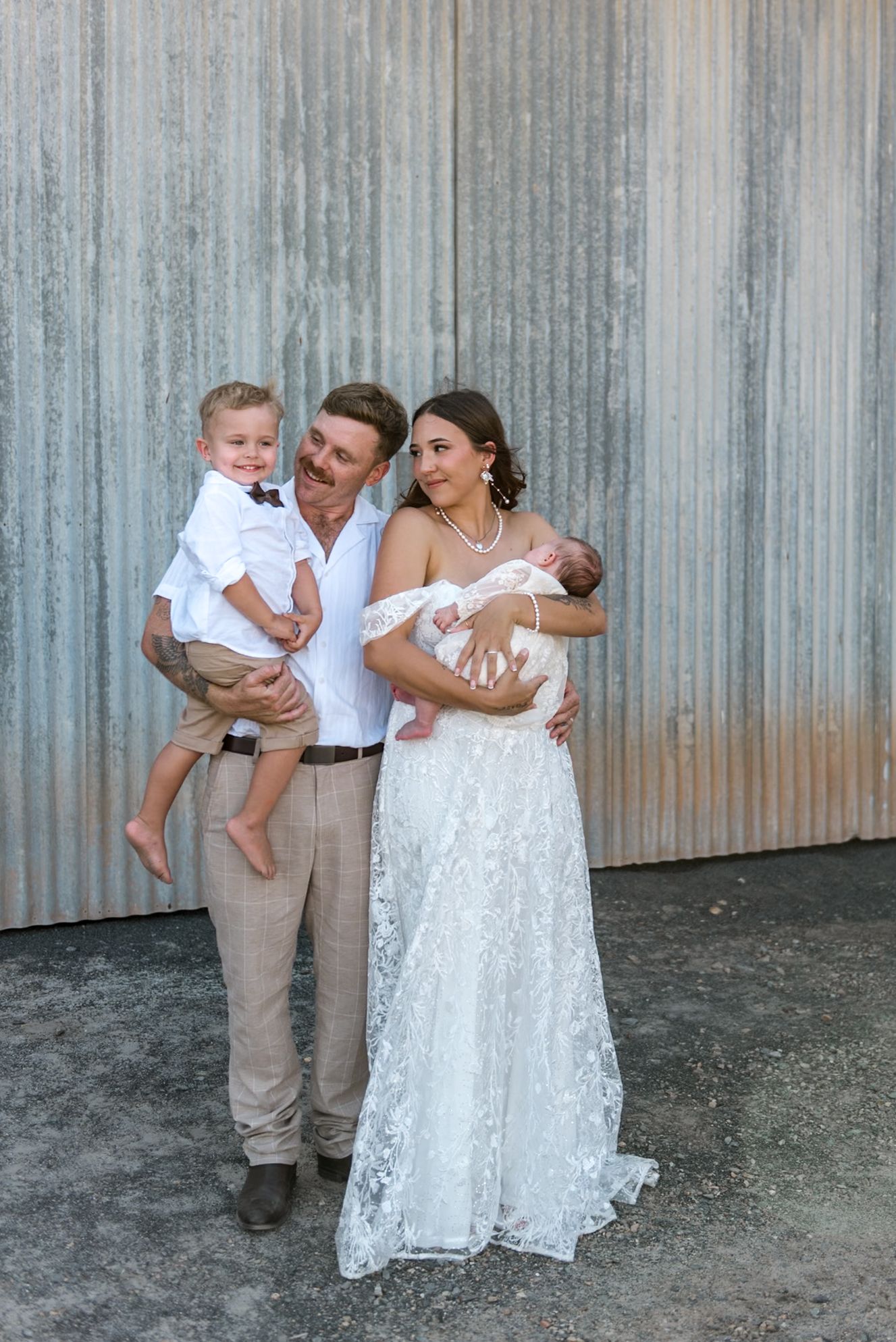 Family portrait with bride in lace gown holding newborn and groom carrying toddler son against rustic metal wall.