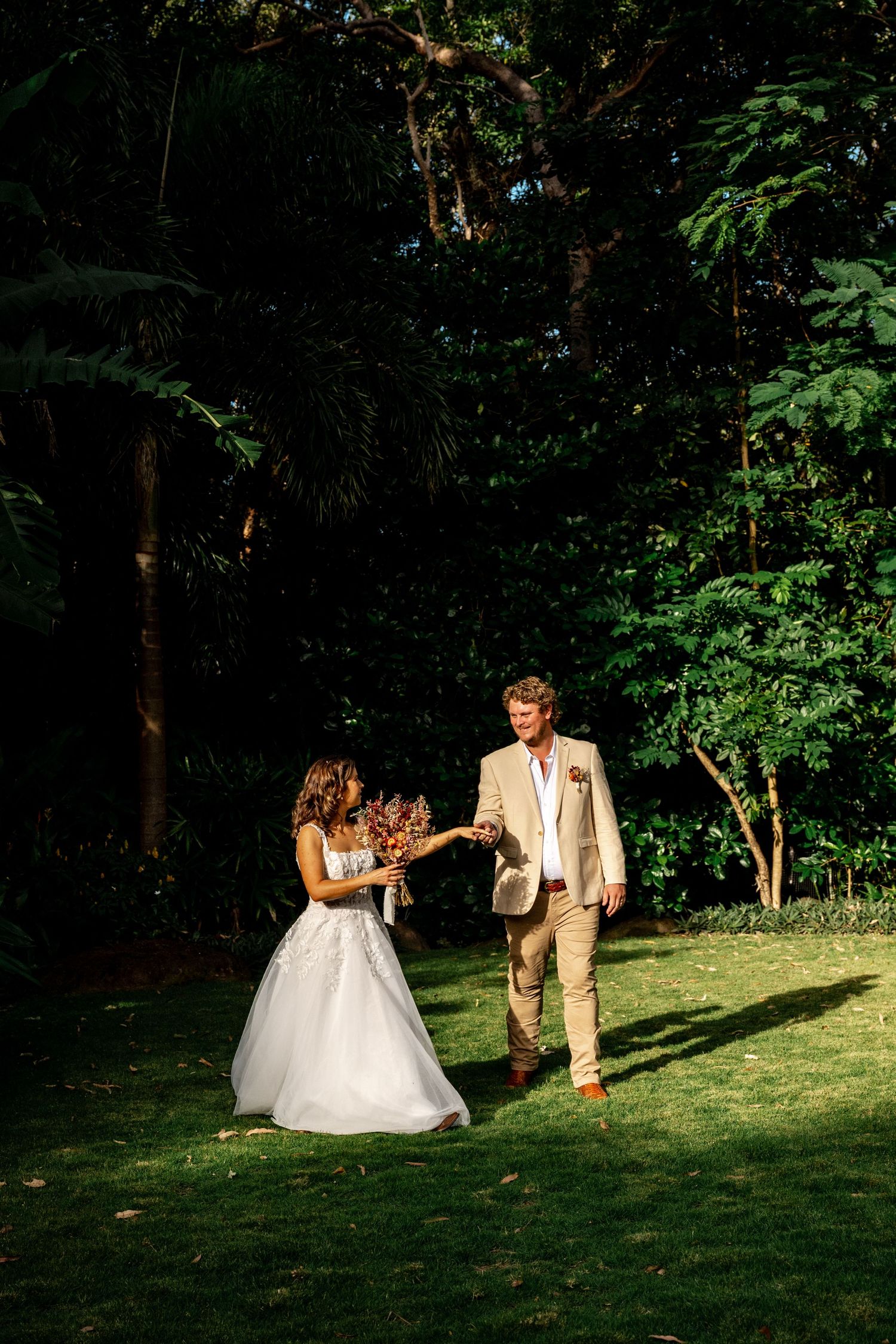 Joyful newlyweds dance playfully on green lawn surrounded by lush tropical forest after wedding.