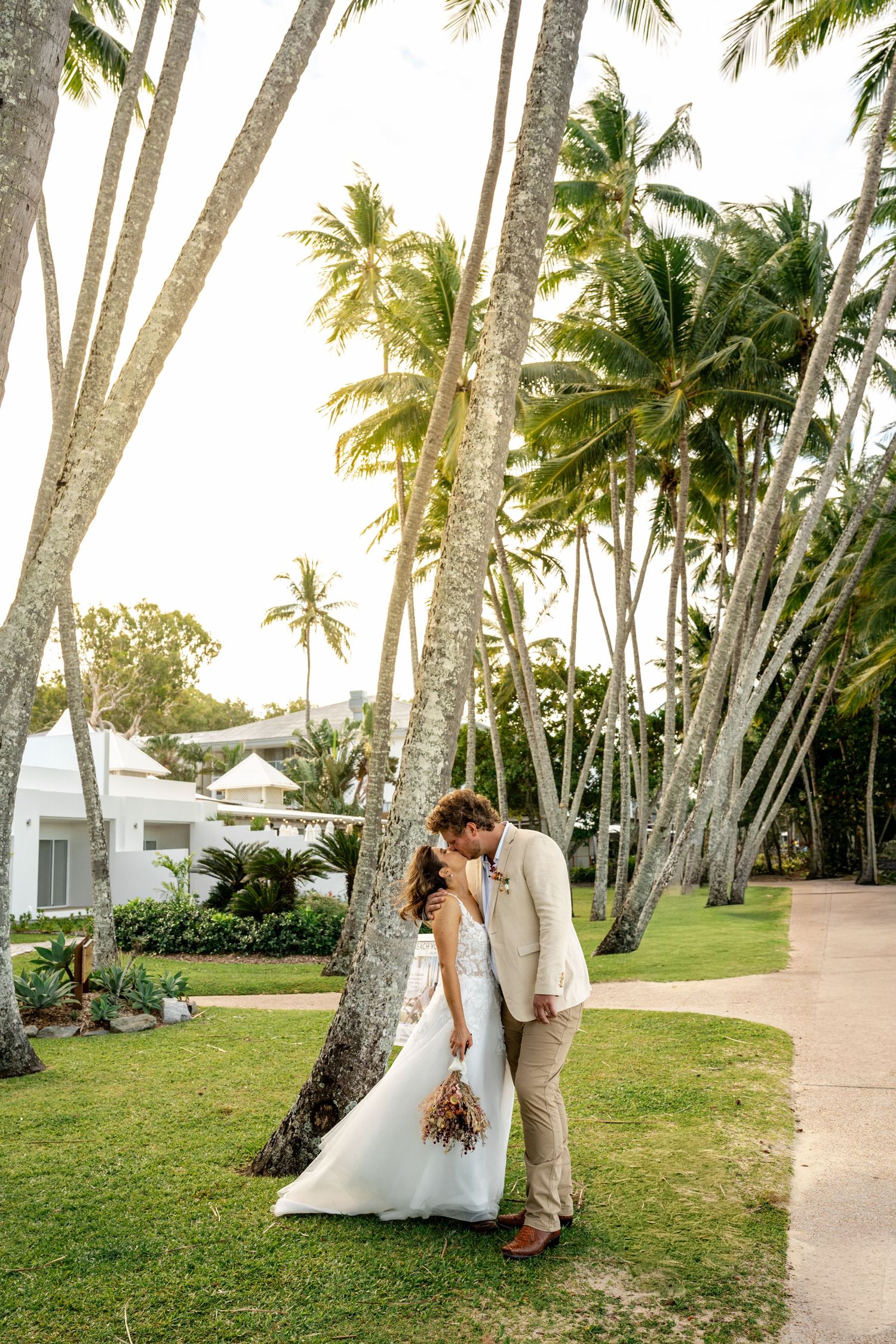 Newlywed couple sharing a romantic kiss among tall palm trees in a lush tropical garden setting.