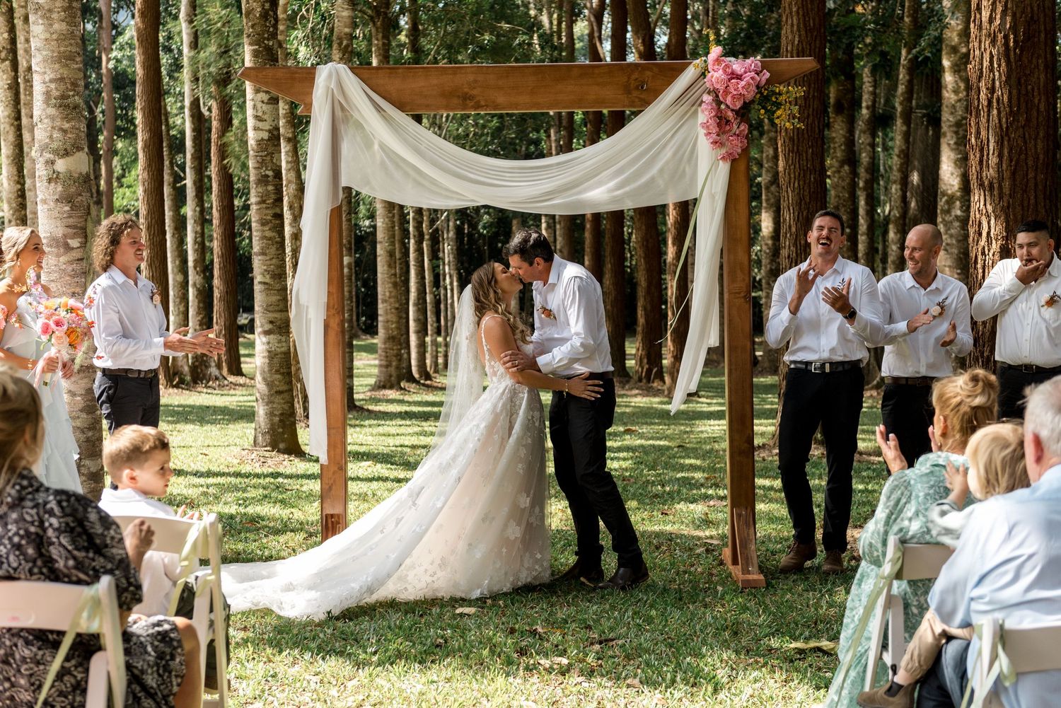 Bride and groom share first kiss under a wooden arch adorned with white draping and pink flowers in a forest.
