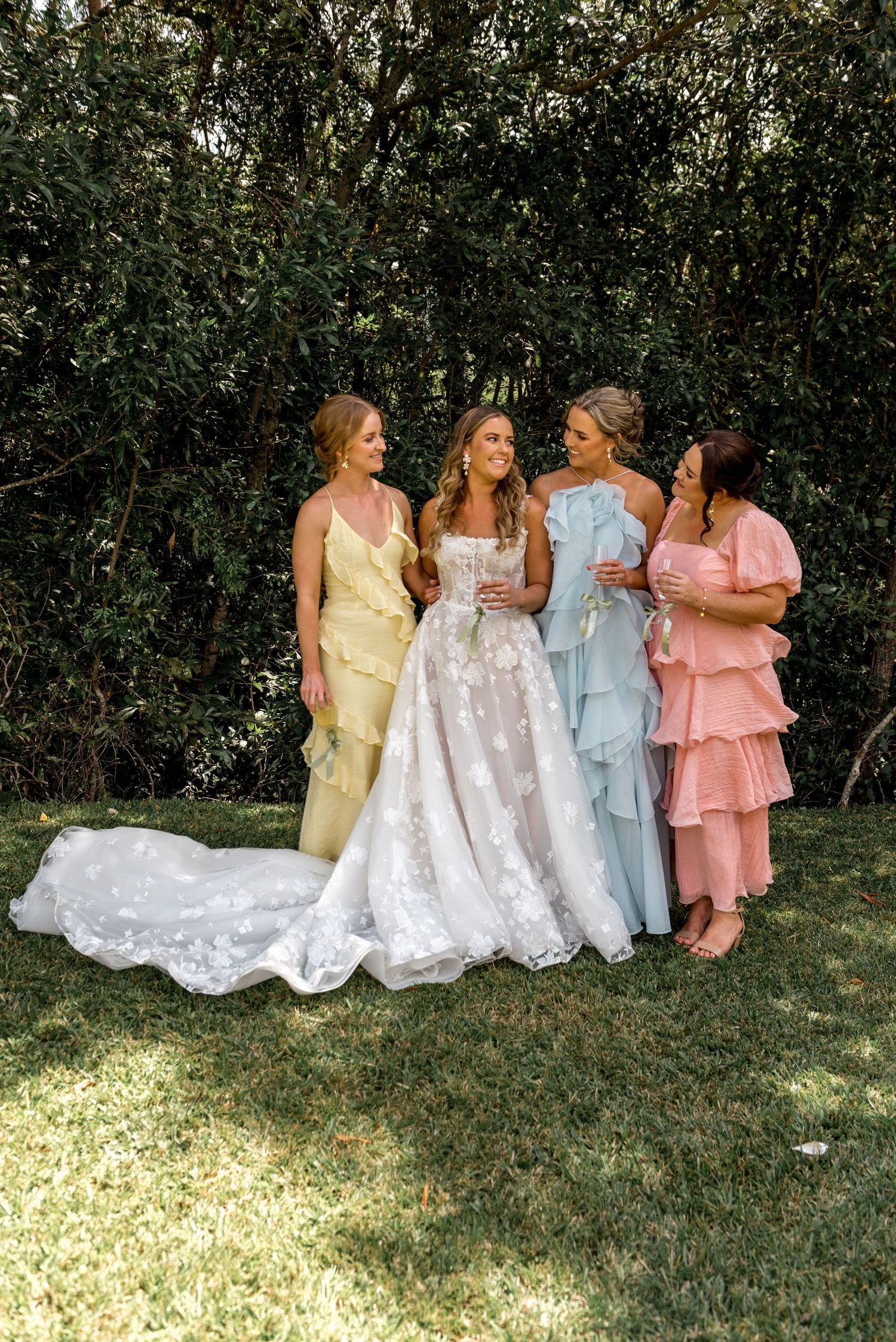 Bride in lace gown shares joyful moment with three bridesmaids in pastel ruffled dresses.