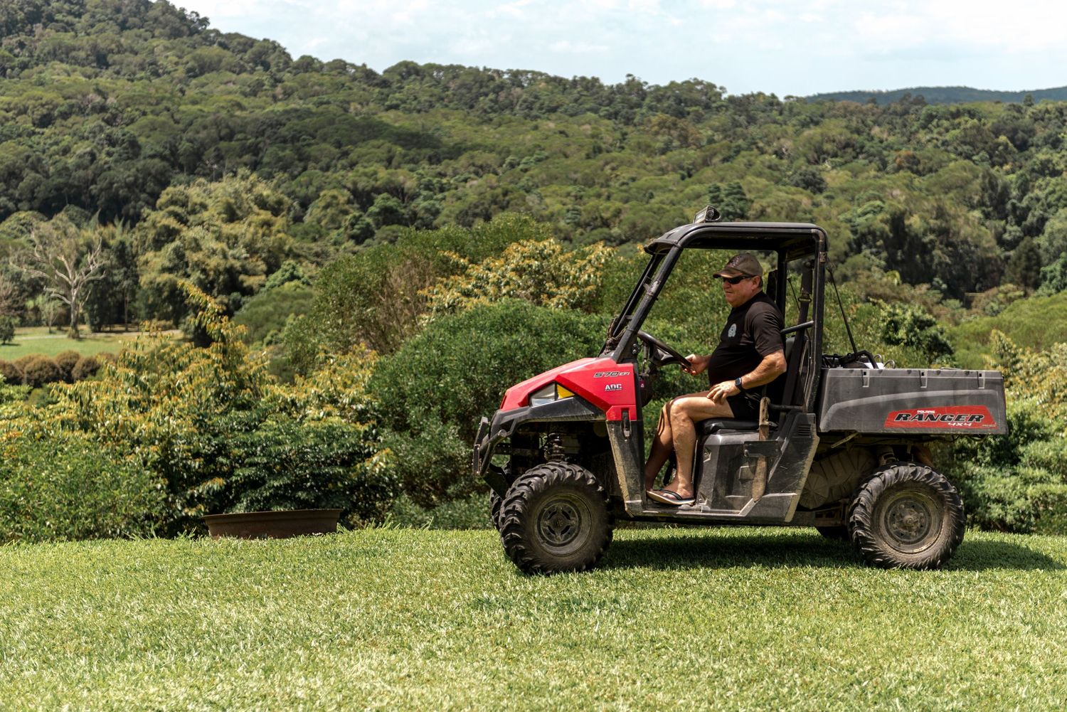 Man driving red utility vehicle across green lawn with forested hills in background.