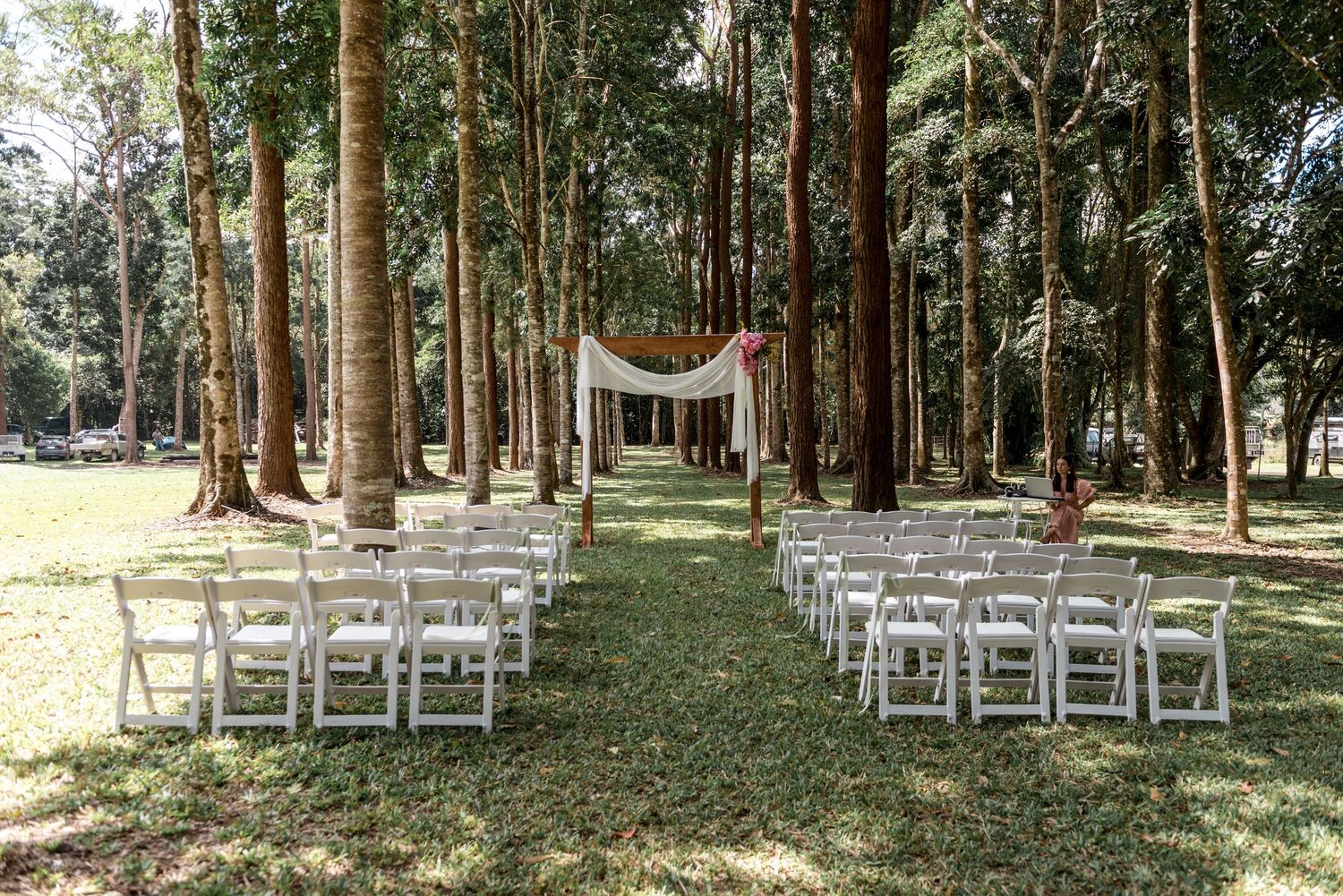 Outdoor wedding ceremony setup with white folding chairs and fabric arch between tall trees in a forest.