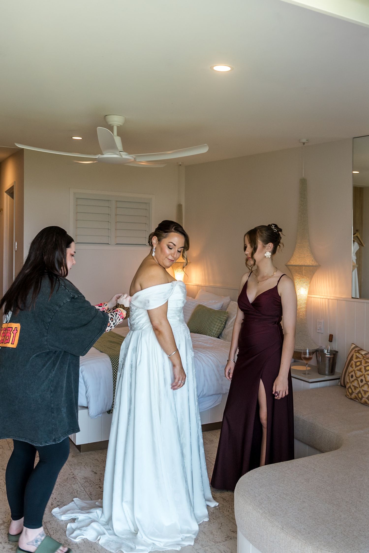 Bride in off-shoulder white gown smiling as two women assist her in a hotel room.