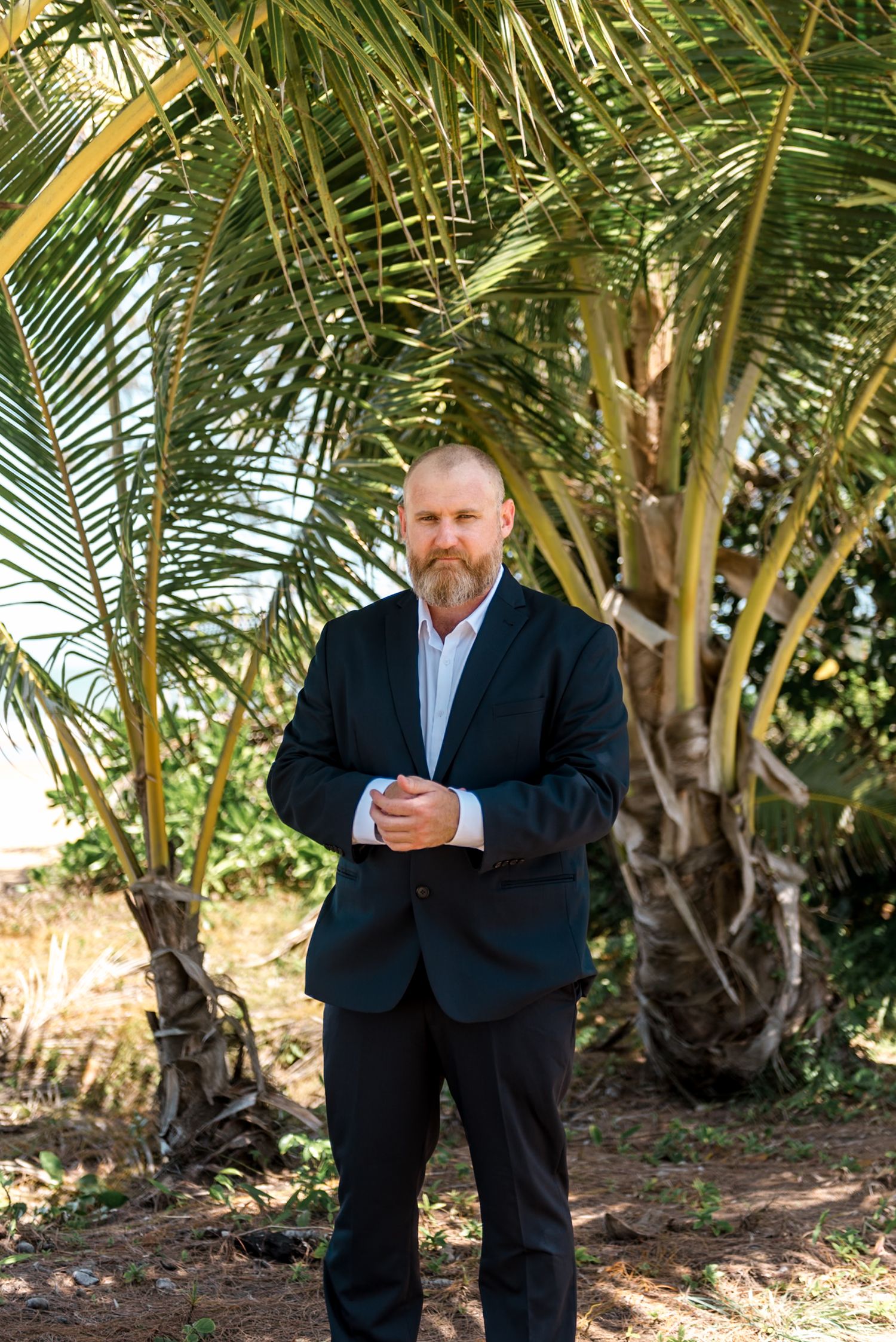 Bearded man in dark suit posing outdoors in front of lush green palm trees on a sunny day.