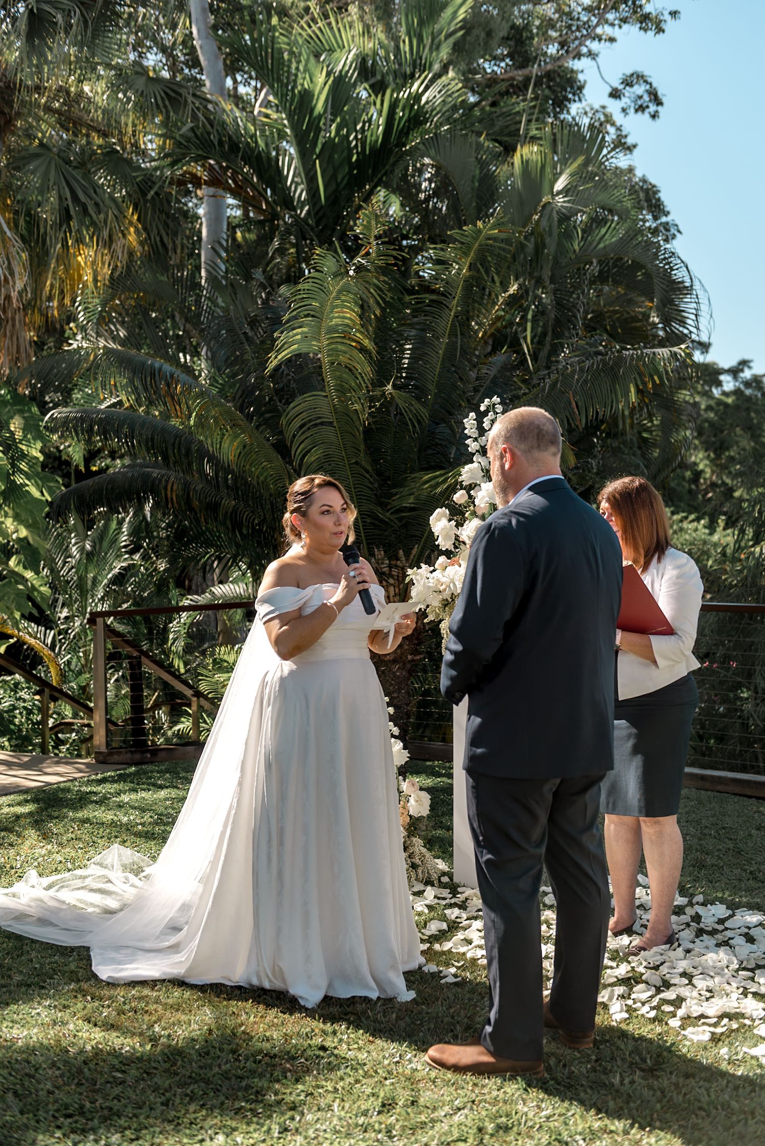 Bride reads wedding vows to groom as officiant holds red book during outdoor tropical garden ceremony.