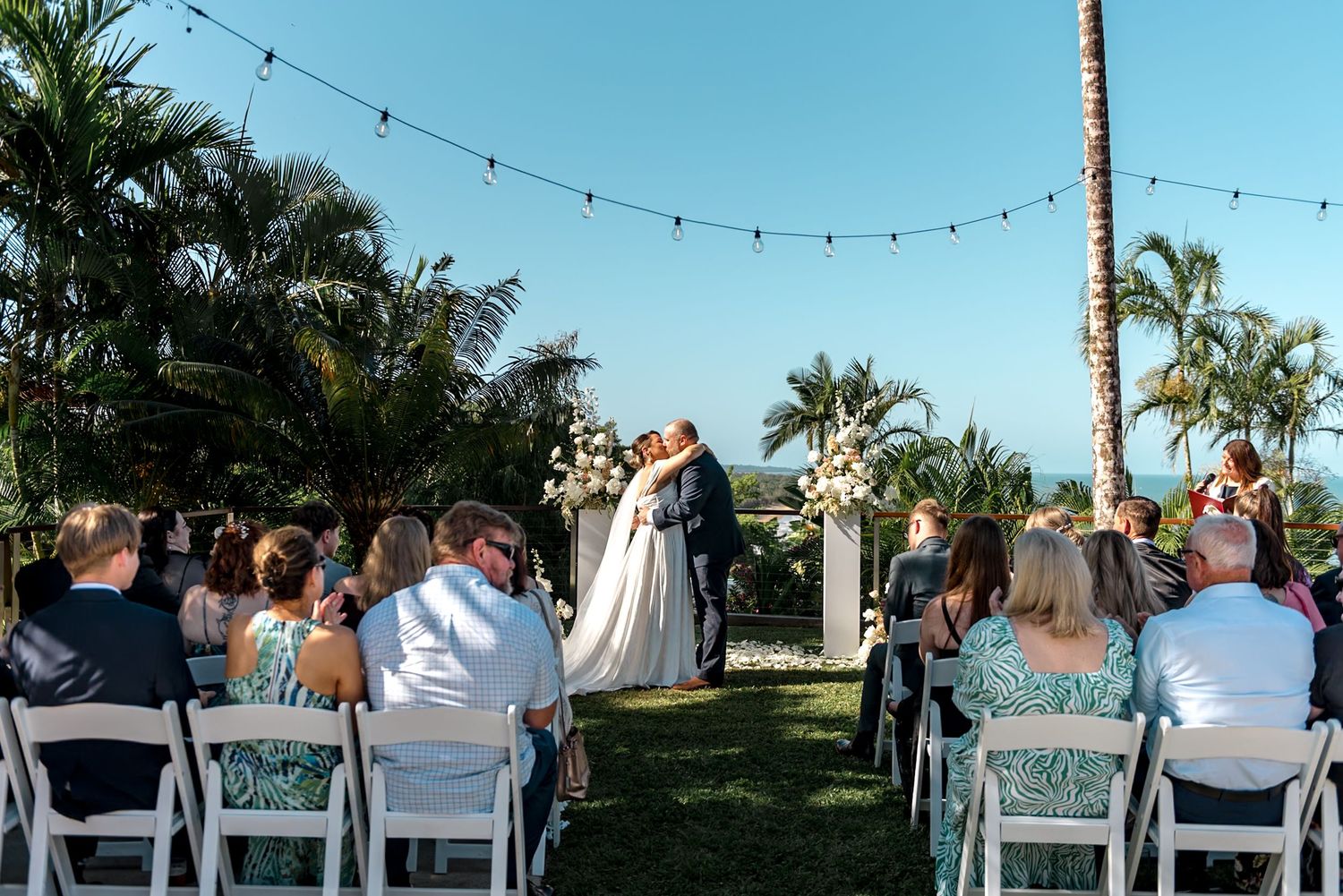 Couple sharing first kiss at outdoor wedding ceremony with tropical palms and string lights.