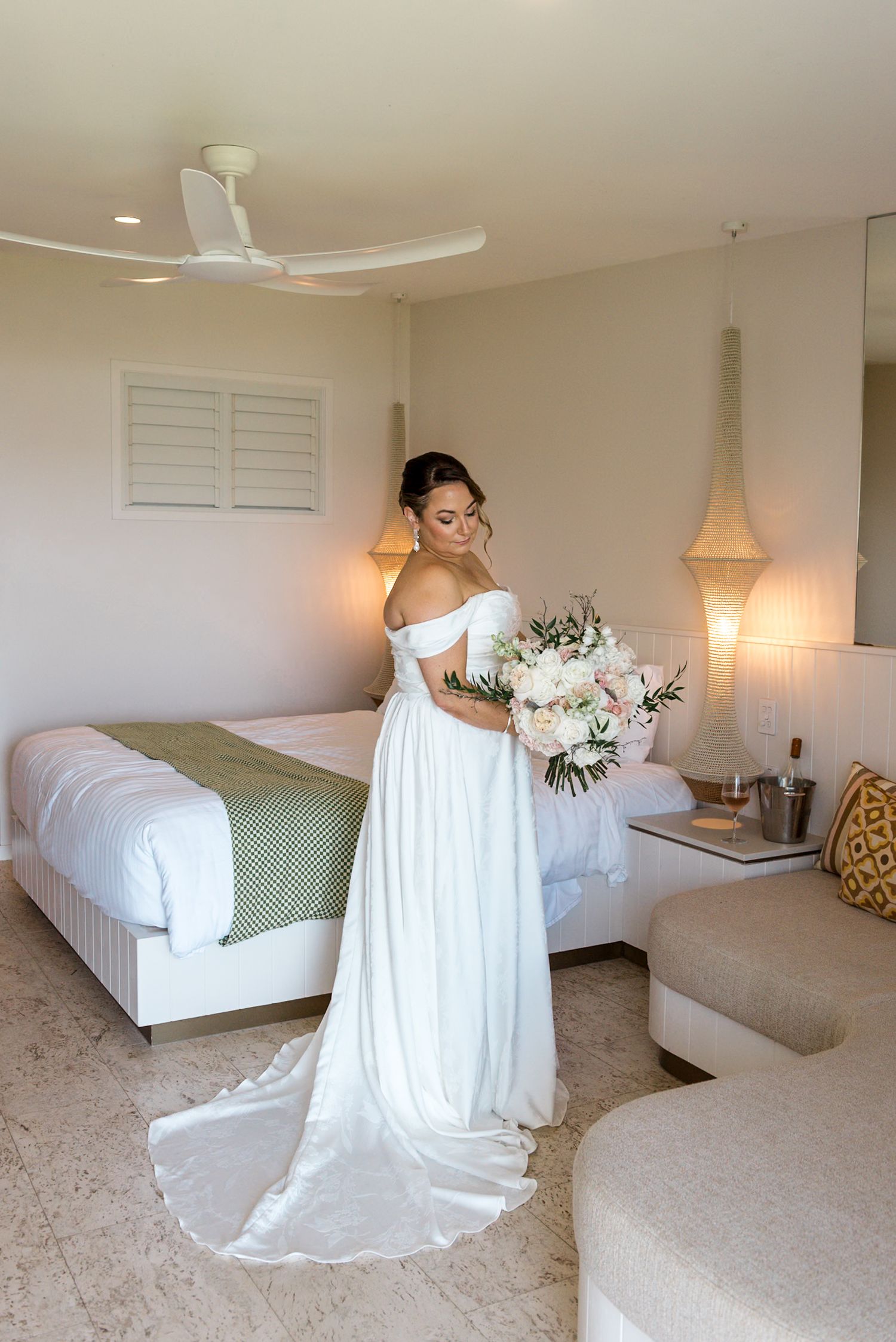 Bride in elegant off-shoulder white wedding dress with long train holding a floral bouquet in a hotel suite.