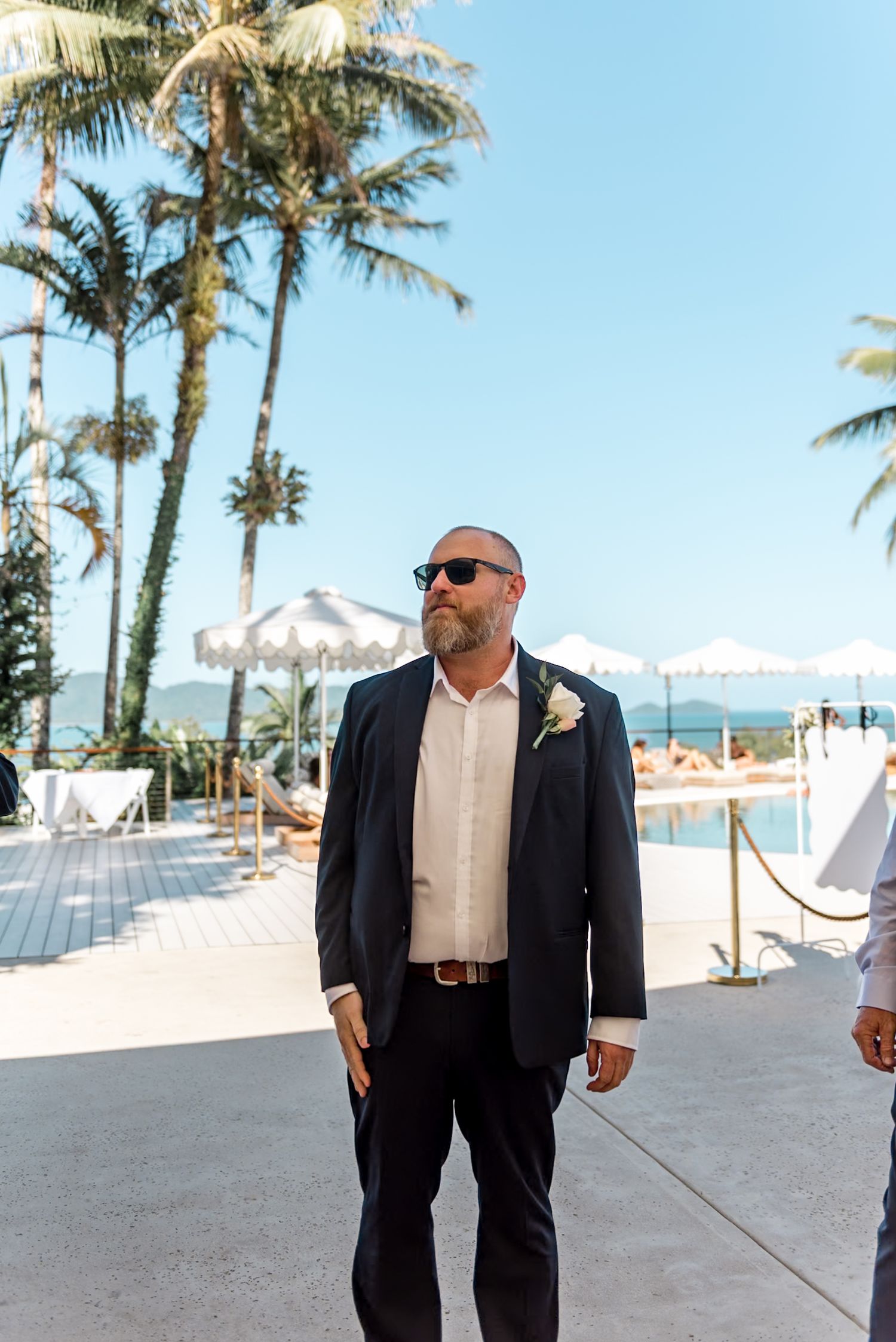 Bald groom in navy suit with boutonniere stands by tropical poolside wedding venue with palm trees.