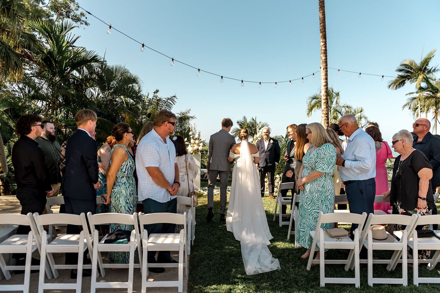 Bride walks down aisle at outdoor tropical wedding ceremony with guests standing and white chairs lining the path.
