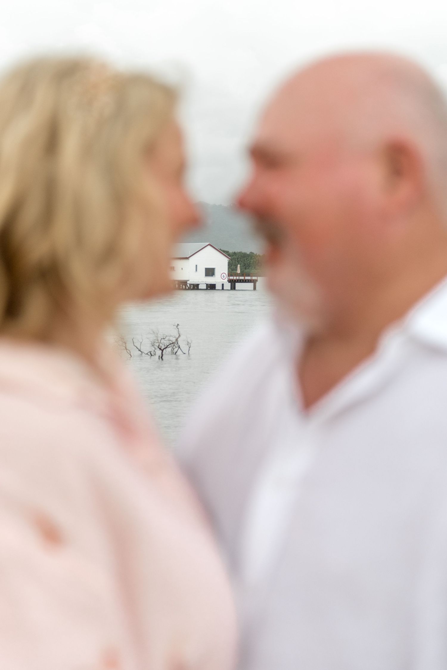 Blurred couple almost kissing with a white waterfront boathouse in the background.