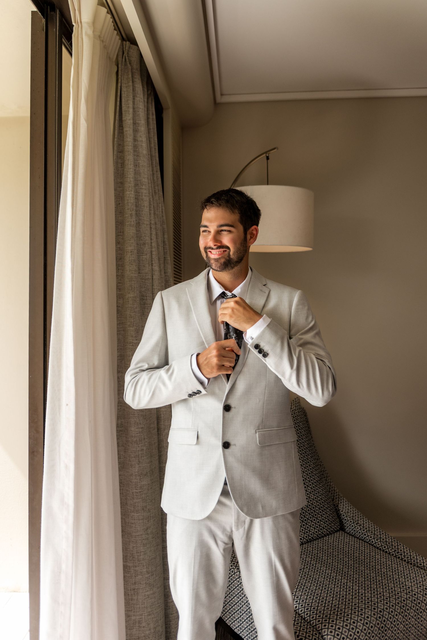 Happy groom in light gray suit smiling while adjusting his dark tie, standing near a window in a hotel room.