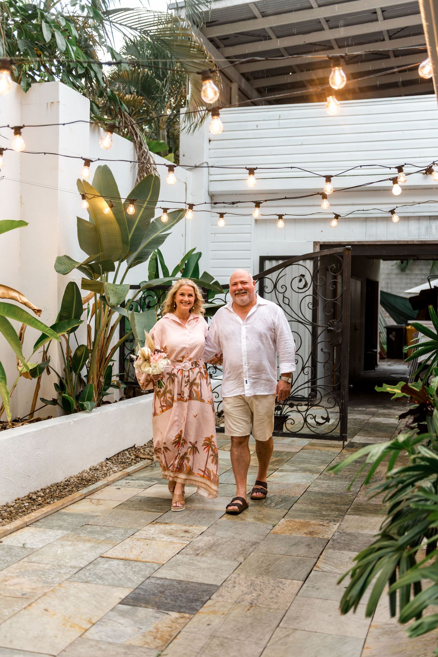 Happy bride and groom walking hand in hand through tropical courtyard adorned with warm festoon string lights.