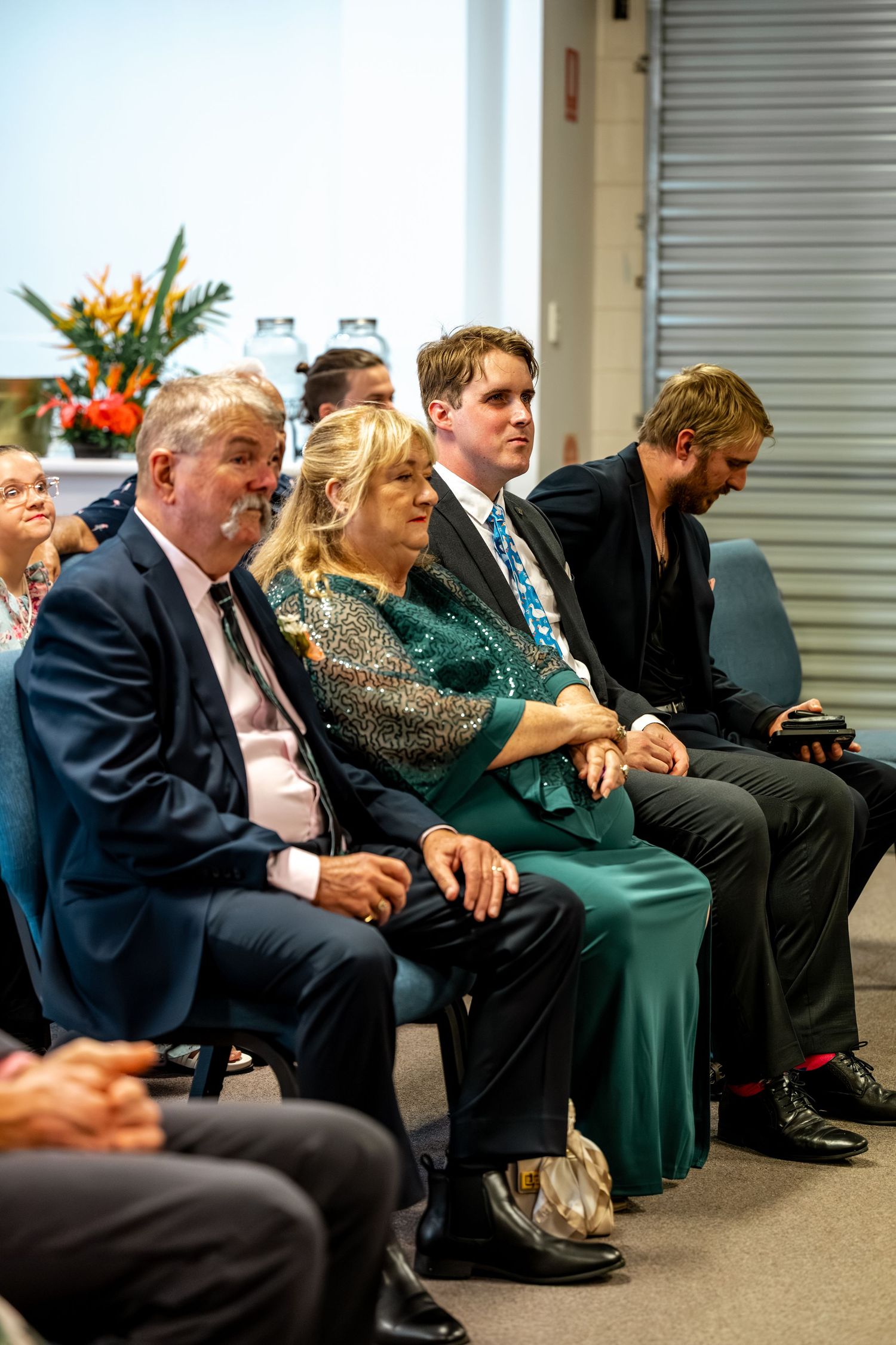 Wedding guests seated in ceremony including couple in navy suit and teal sequin dress holding hands in front row.