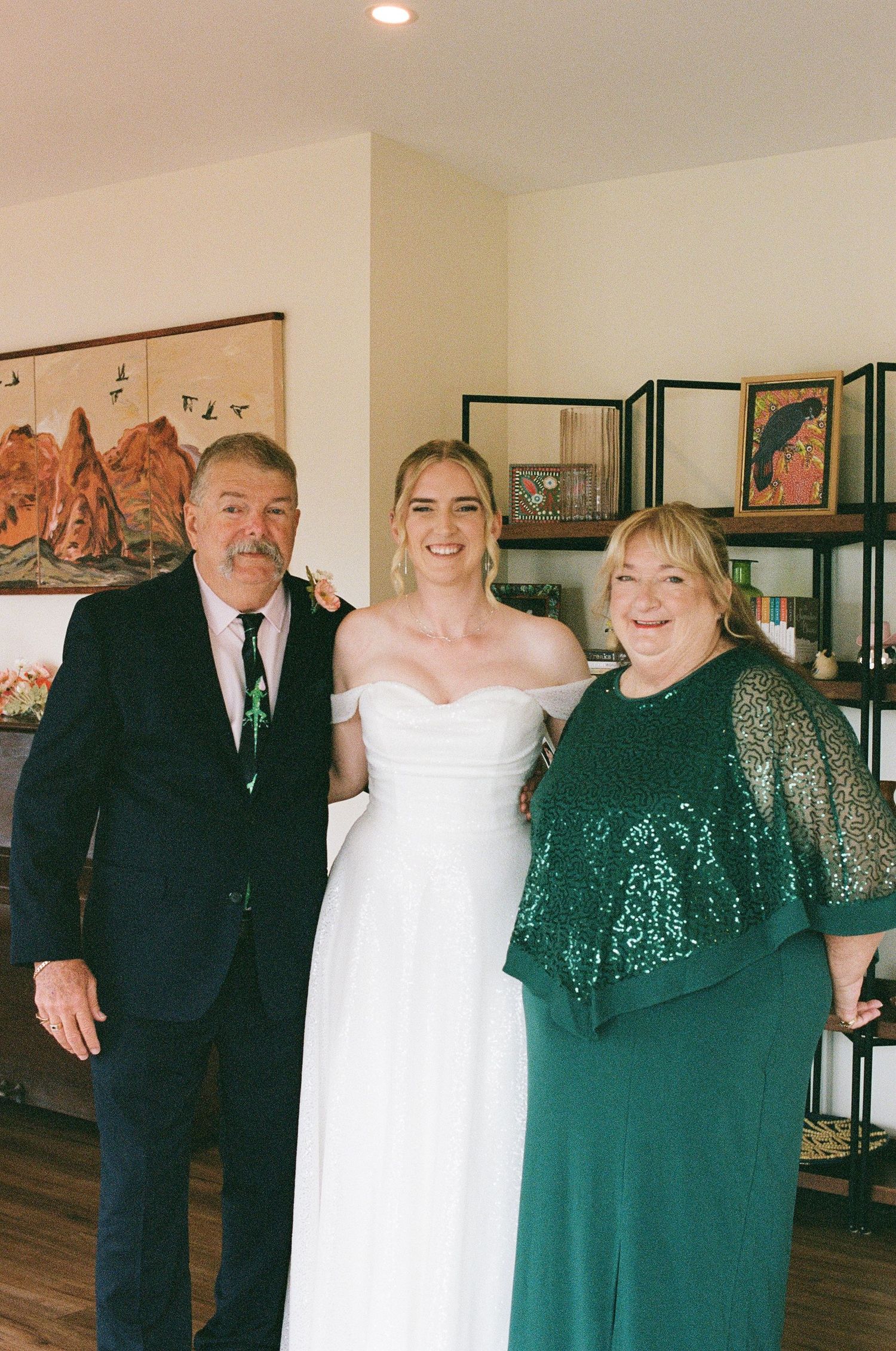 Bride in white off-shoulder gown posing with parents at wedding venue before ceremony.