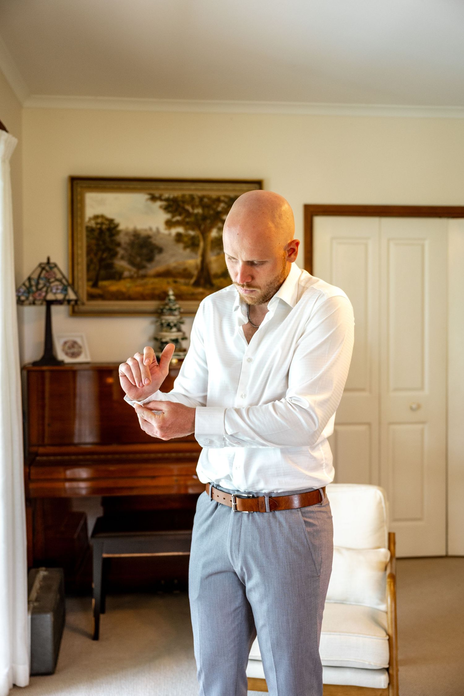 Bald groom in white shirt fastening cufflinks while getting ready for wedding day.