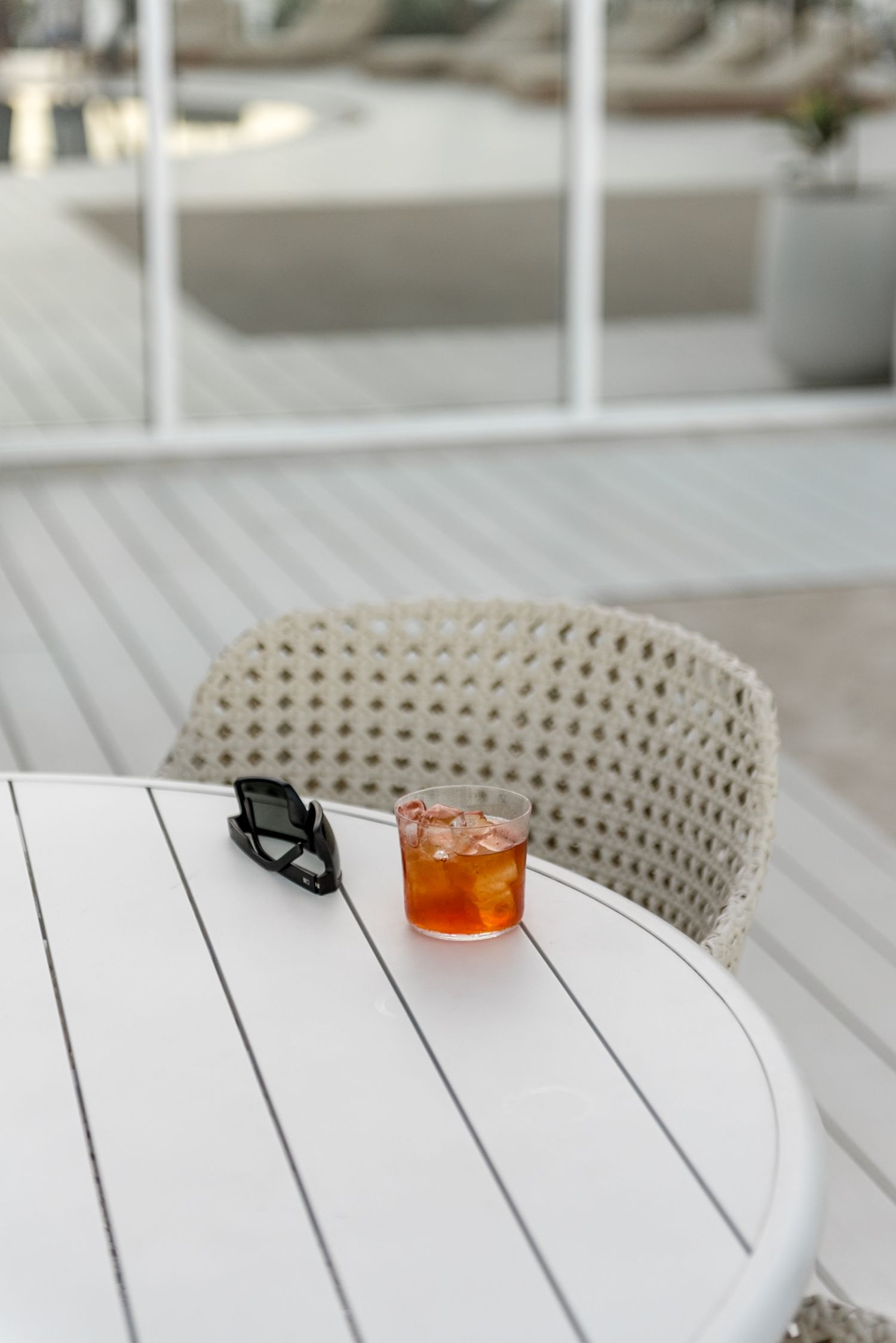 Cocktail drink with ice in a rocks glass beside sunglasses on a white outdoor table at a luxury resort.