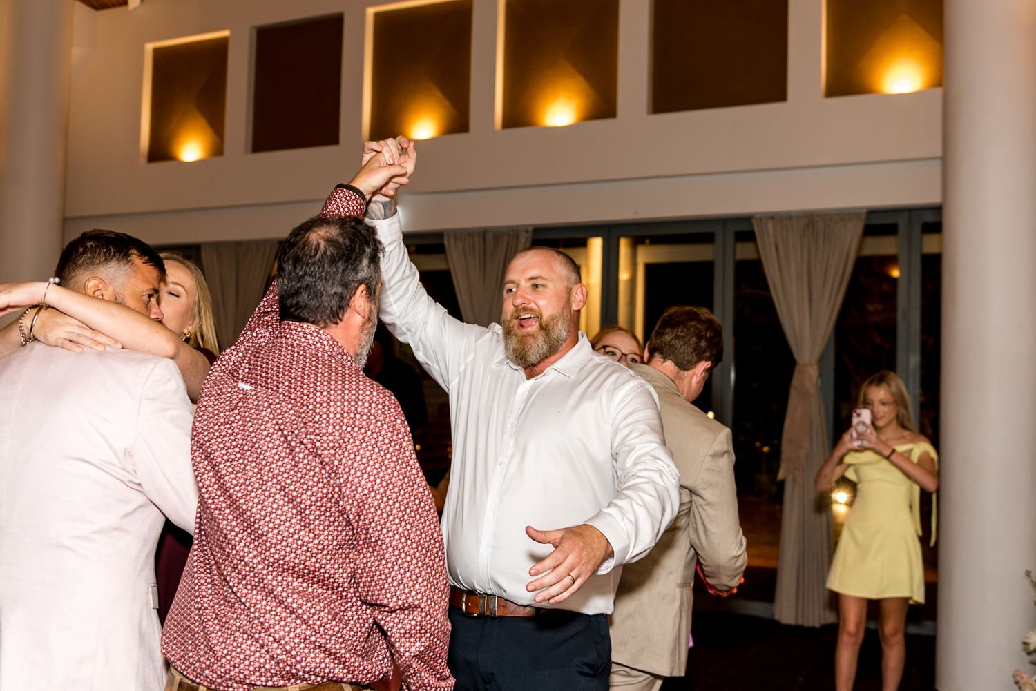 Guests dance energetically at a wedding reception in a warmly lit venue.