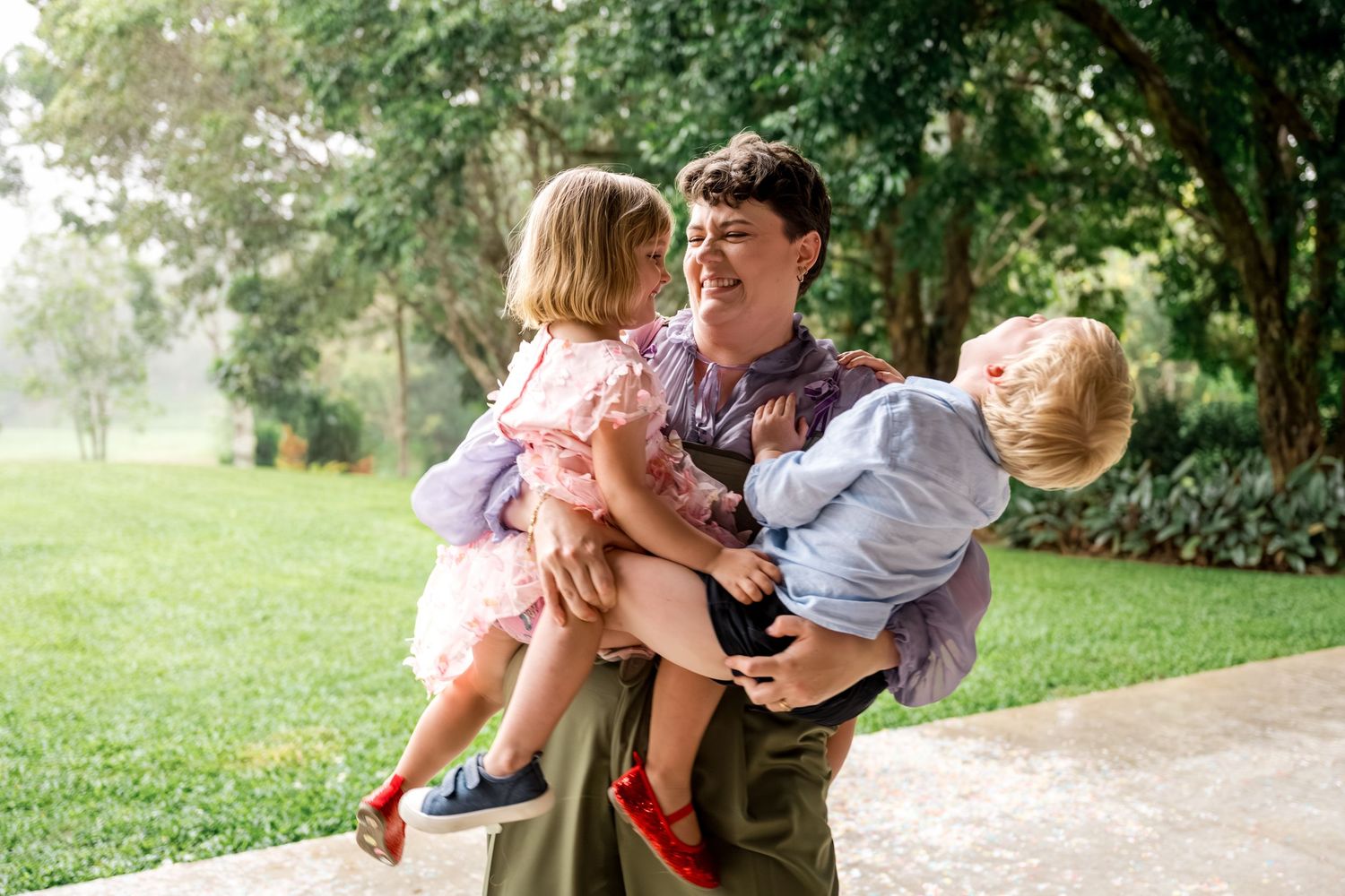 Smiling person holds two young children in lush green garden at outdoor wedding.