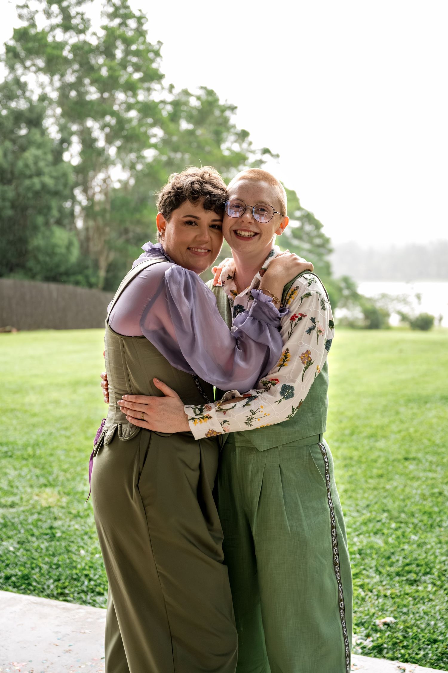 Two women in matching green overalls embrace and smile outdoors on a green lawn.