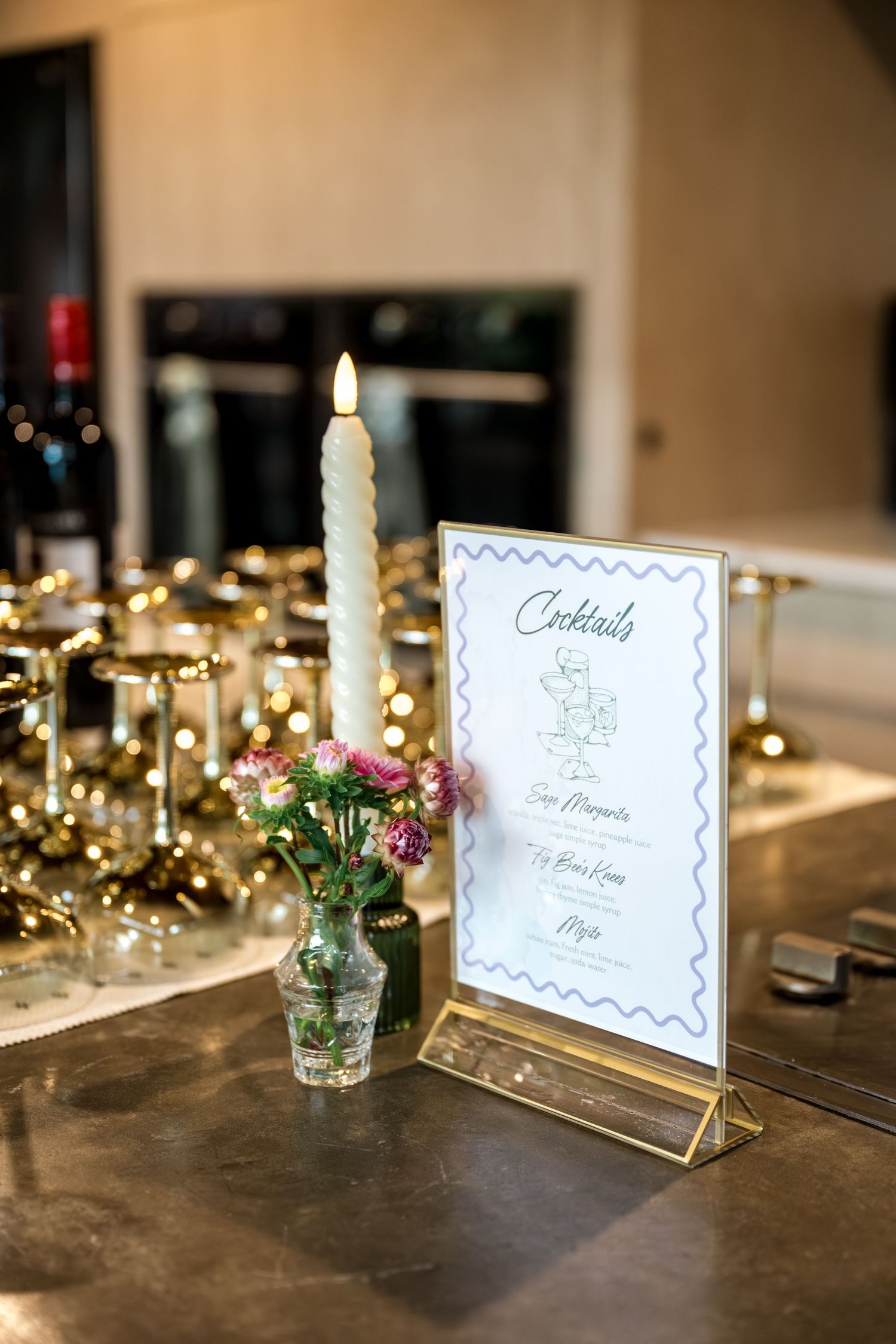 Wedding cocktail bar with lit taper candle, small floral vase, cocktail menu, and gold glassware.