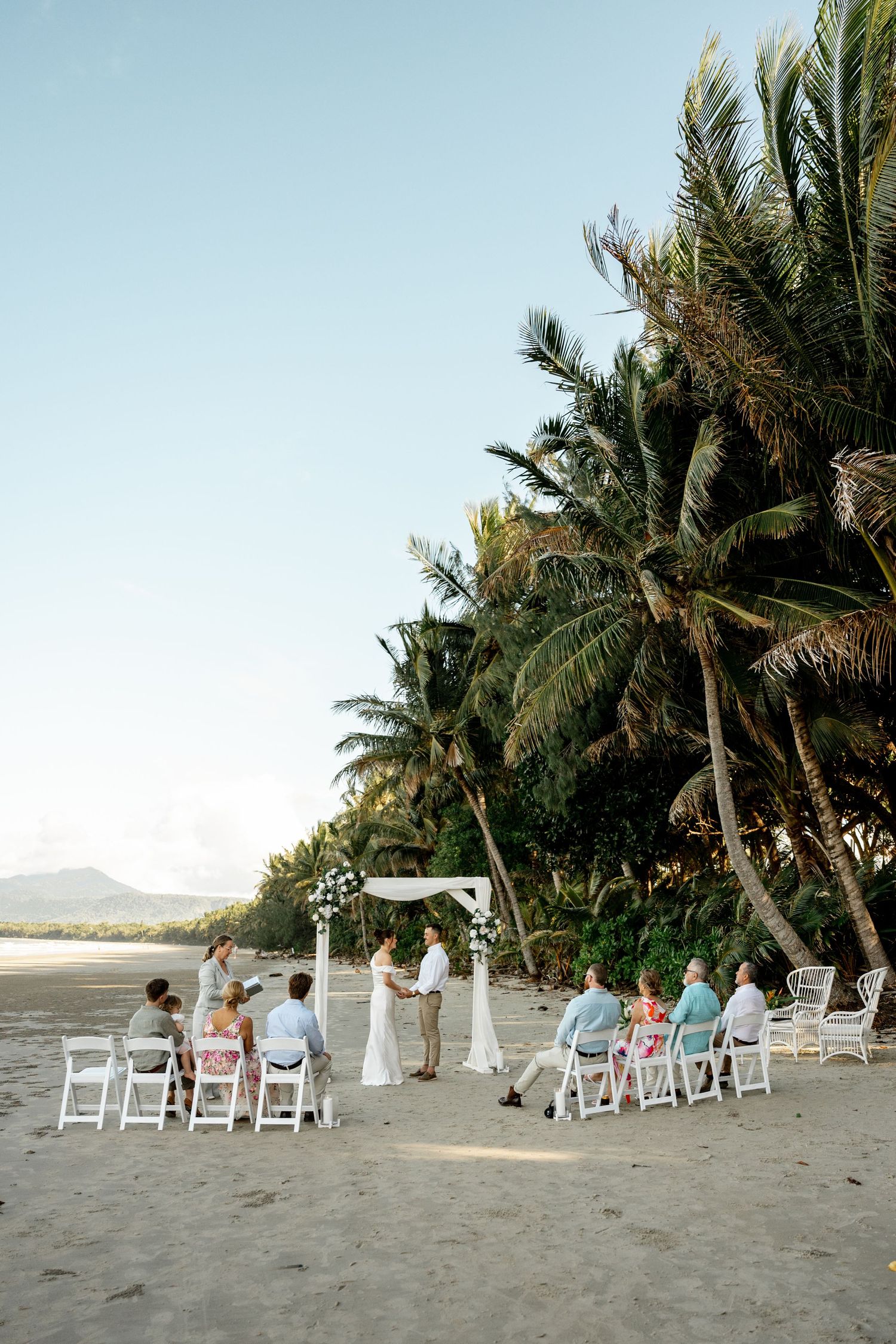 Scenic beach wedding ceremony at sunset with white chairs, floral arch, and tropical palm trees against blue sky at Four Mile Beach.