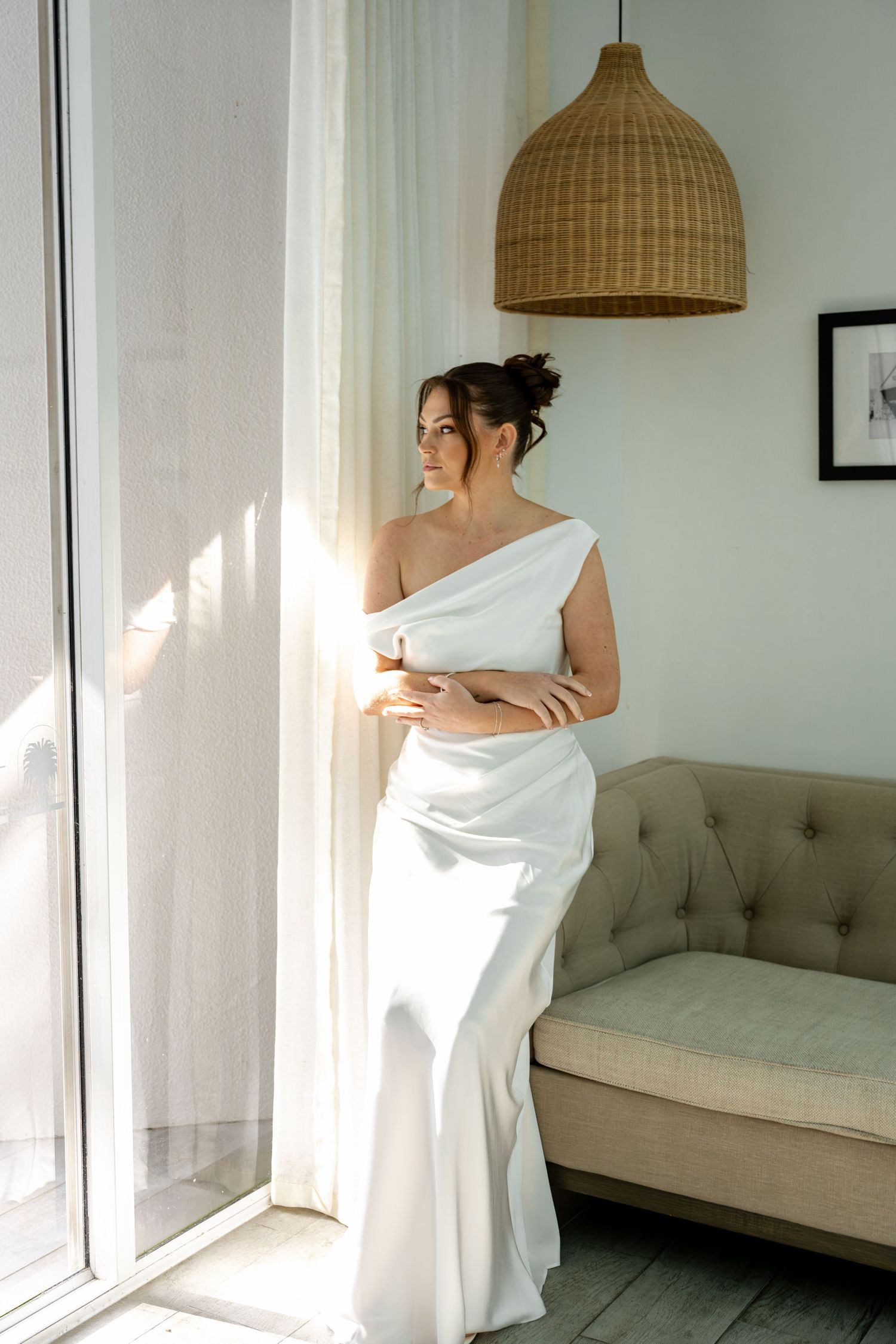 Bride in elegant white one-shoulder gown stands by sunlit window with arms crossed in modern bright room.