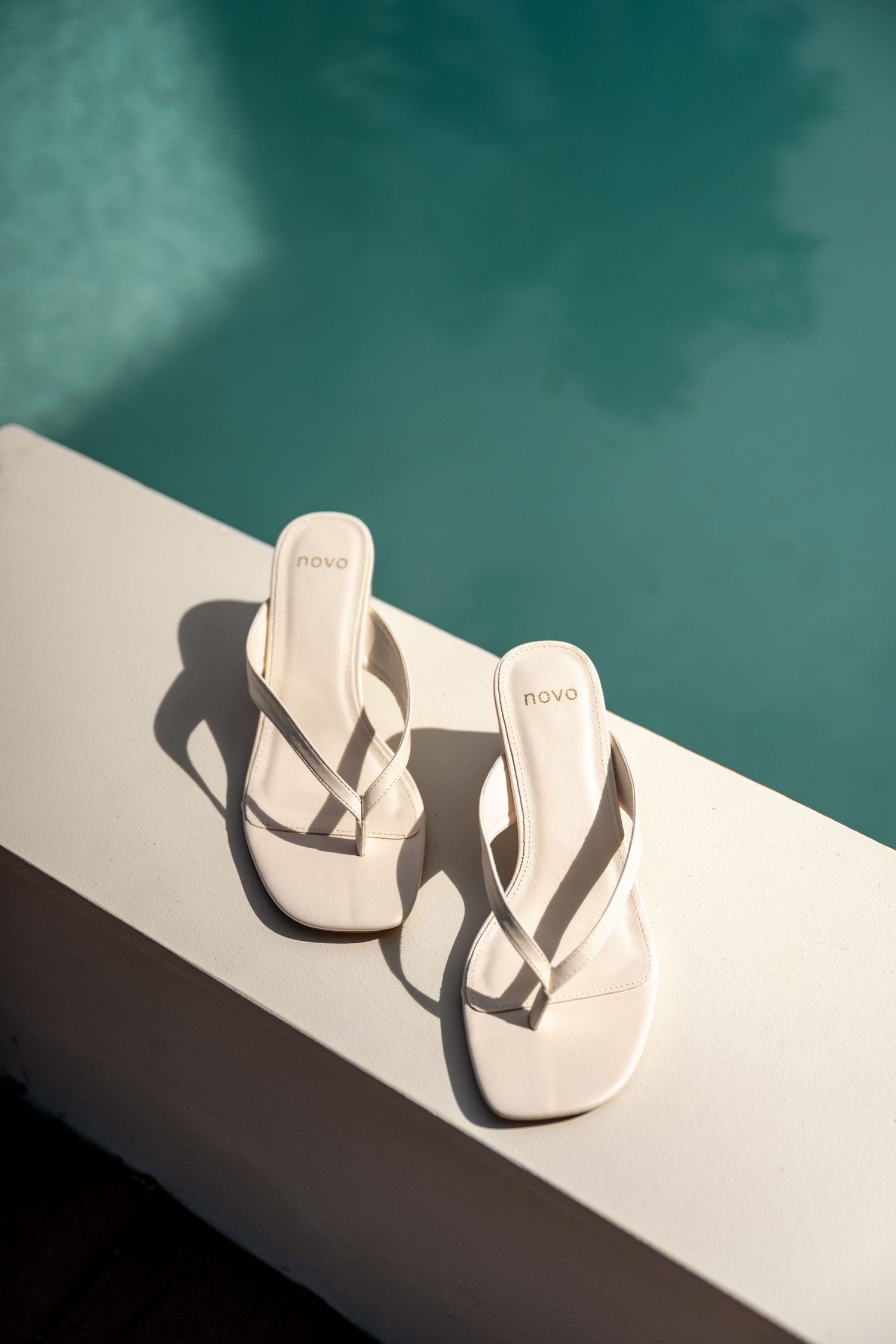 White Novo brand thong sandals placed on edge of turquoise swimming pool in sunlight.