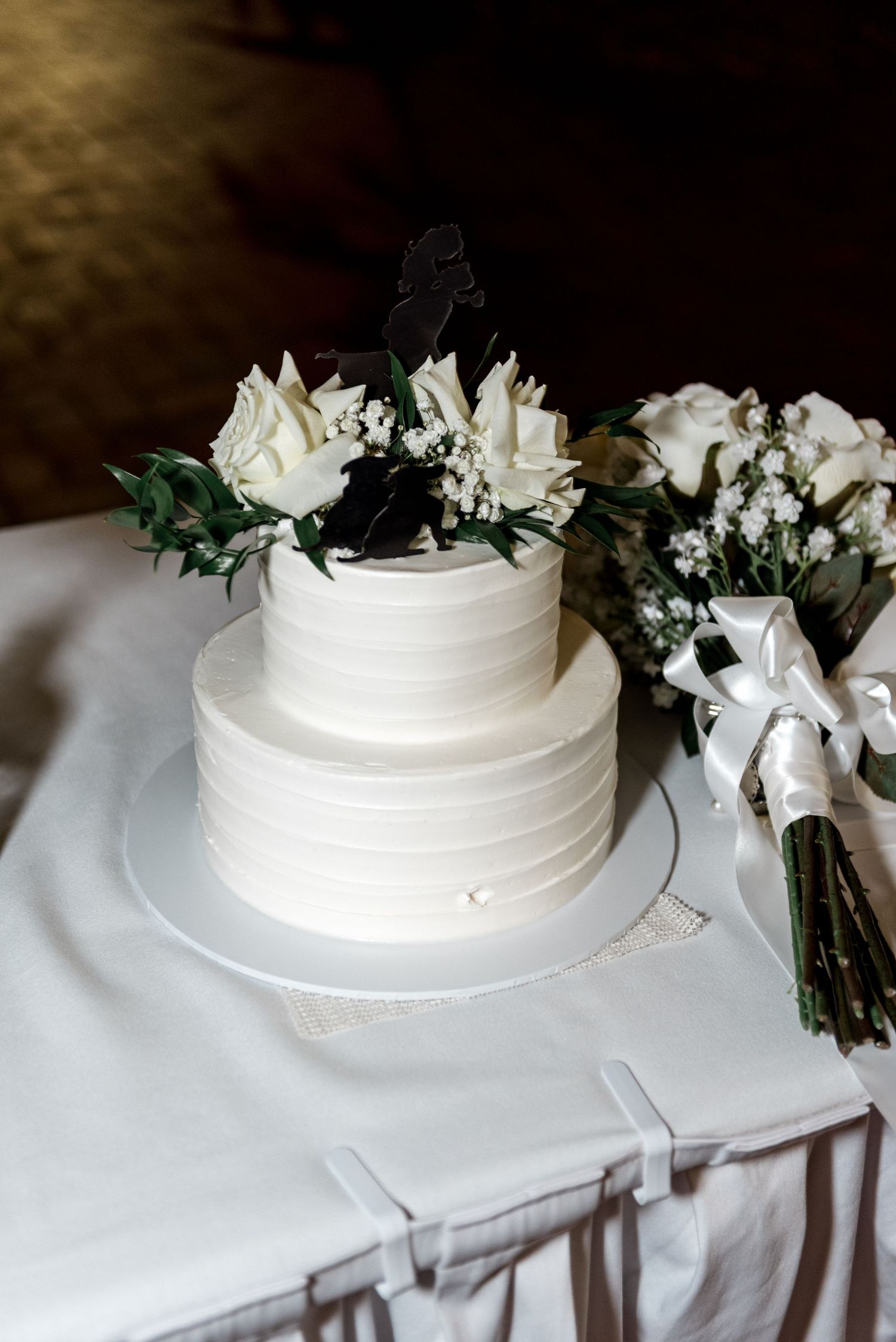 Classic two-tier white ribbed wedding cake topped with white roses and a silhouette cake topper beside a bouquet.