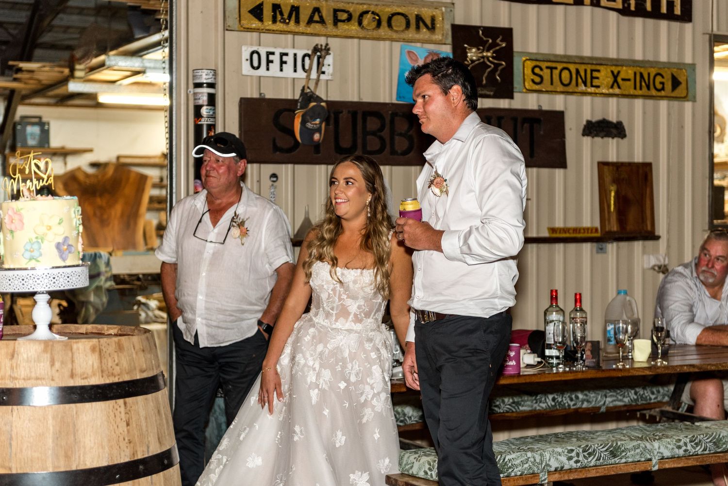 Bride in lace gown standing at rustic bar with wooden barrels and vintage signs while groom speaks at wedding reception.