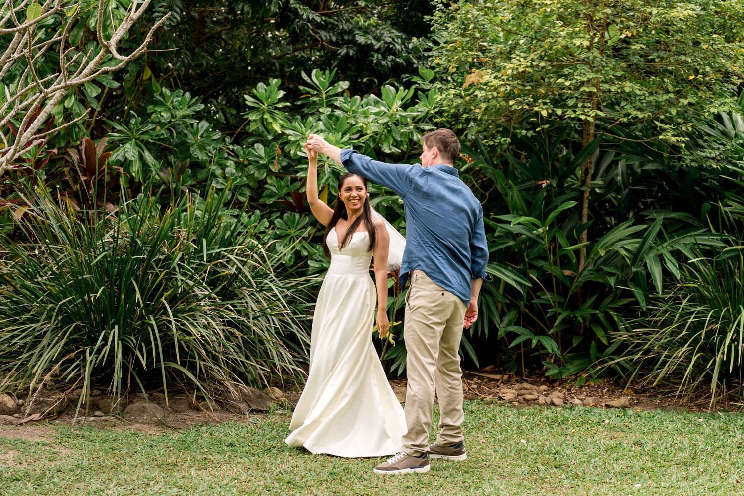 Groom twirling bride in elegant white dress in lush tropical garden after wedding ceremony at Little Cove, Port Douglas.