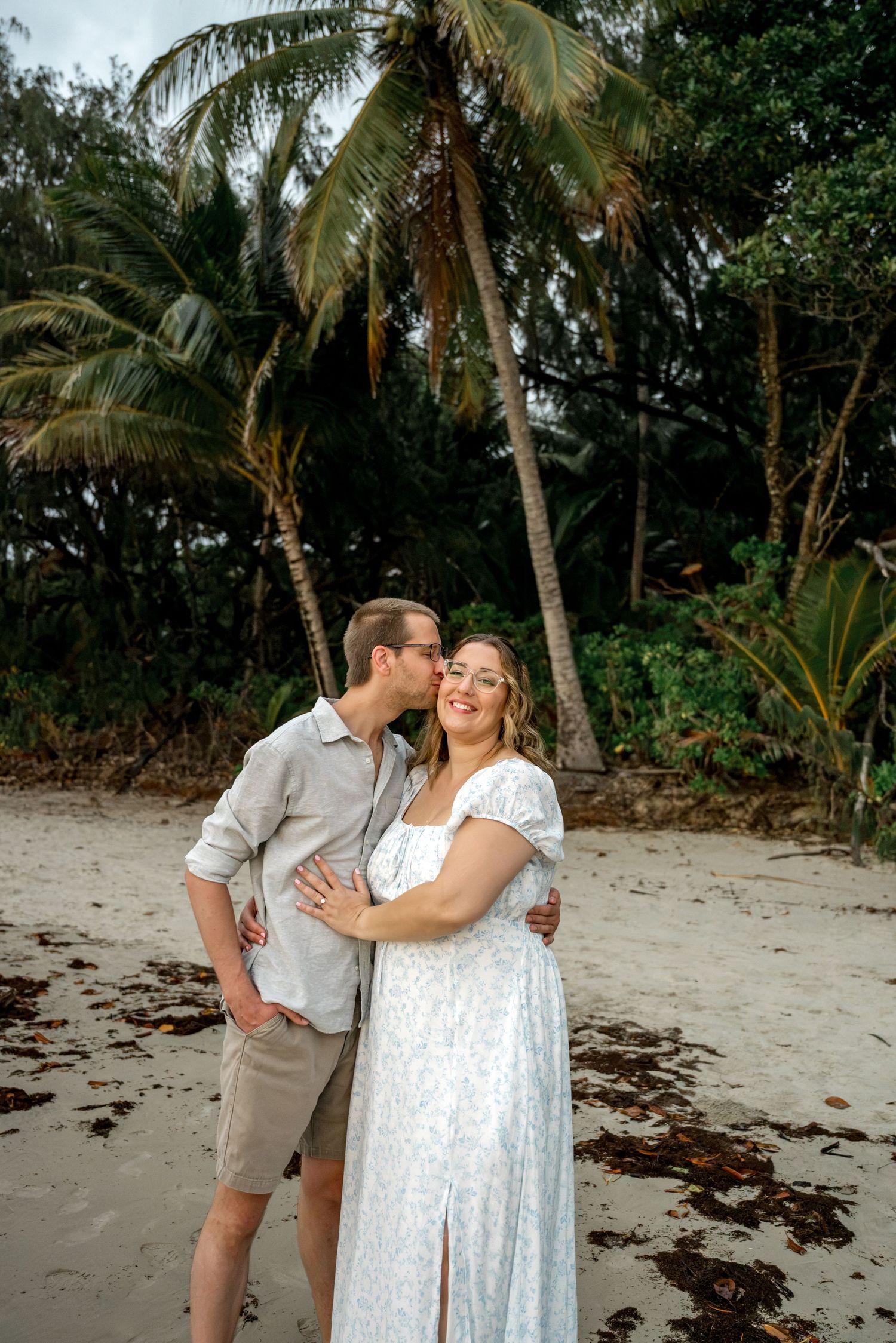 Man kissing smiling woman on cheek while embracing on sandy beach surrounded by tropical palm trees at Four Mile Beach, Port Douglas.