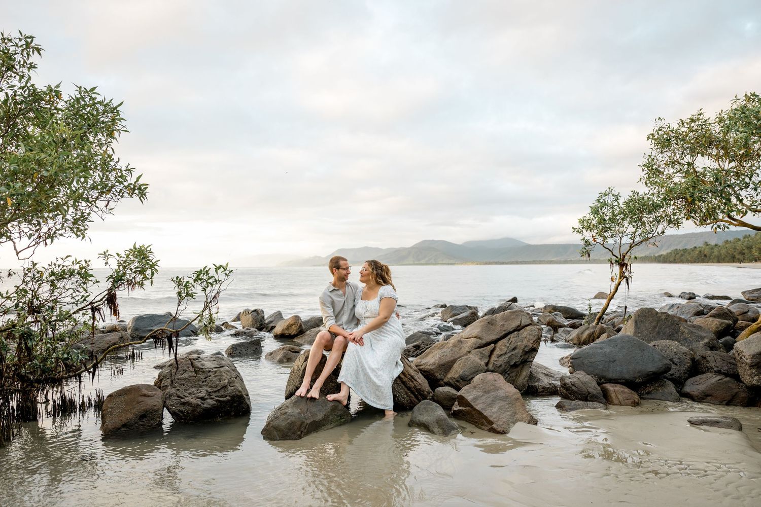 Couple sits together on rocks by calm ocean water surrounded by mangroves under a cloudy sky at Four Mile Beach, Port Douglas.