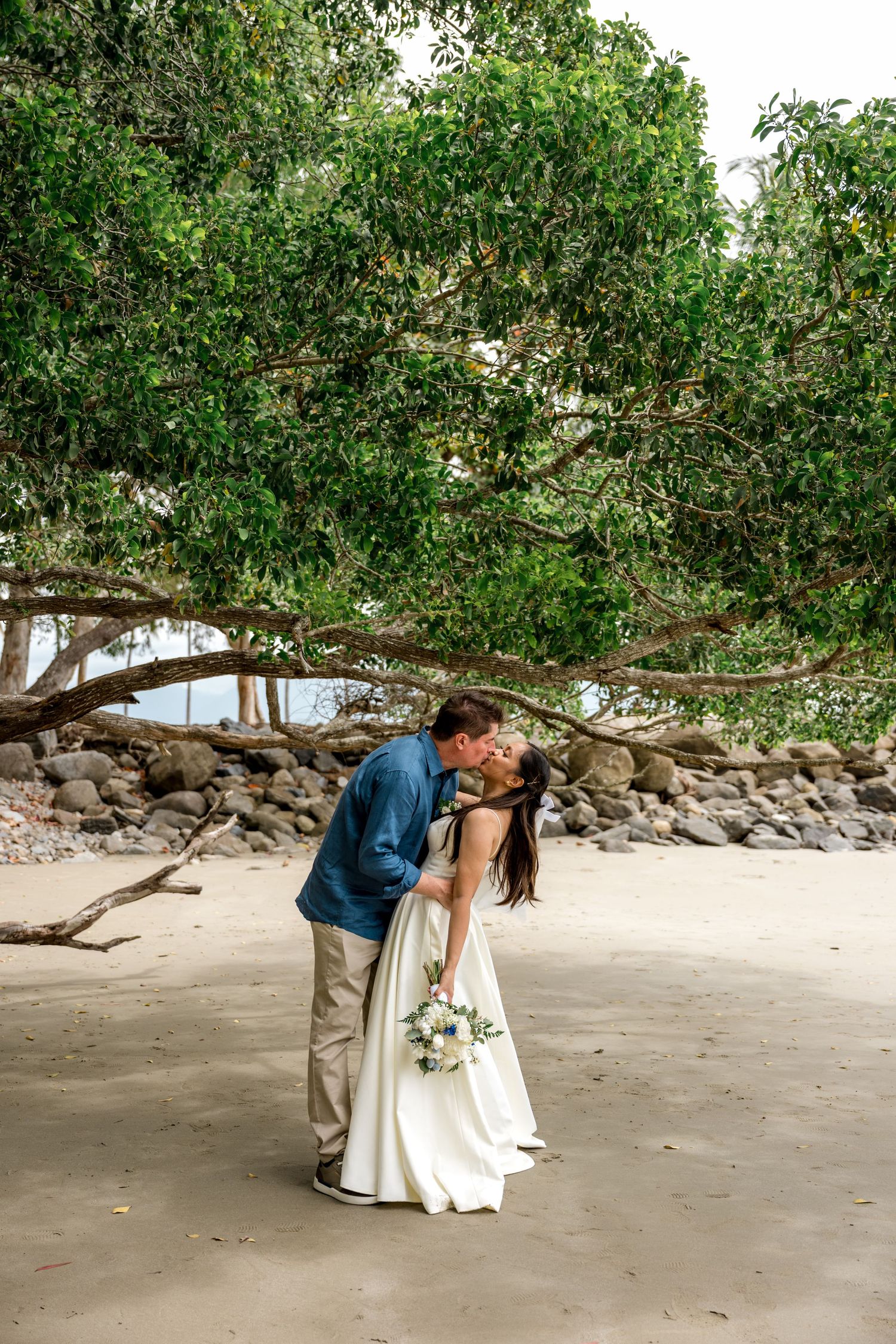 Bride and groom sharing a kiss on a sandy beach beneath a large green tree at Little Cove, Port Douglas.