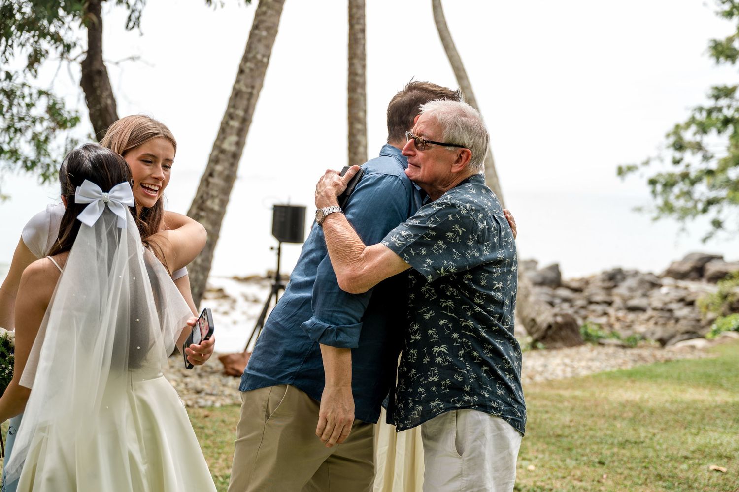 Groom hugging an older male guest while bride smiles at a tropical outdoor wedding at Little Cove, Port Douglas.