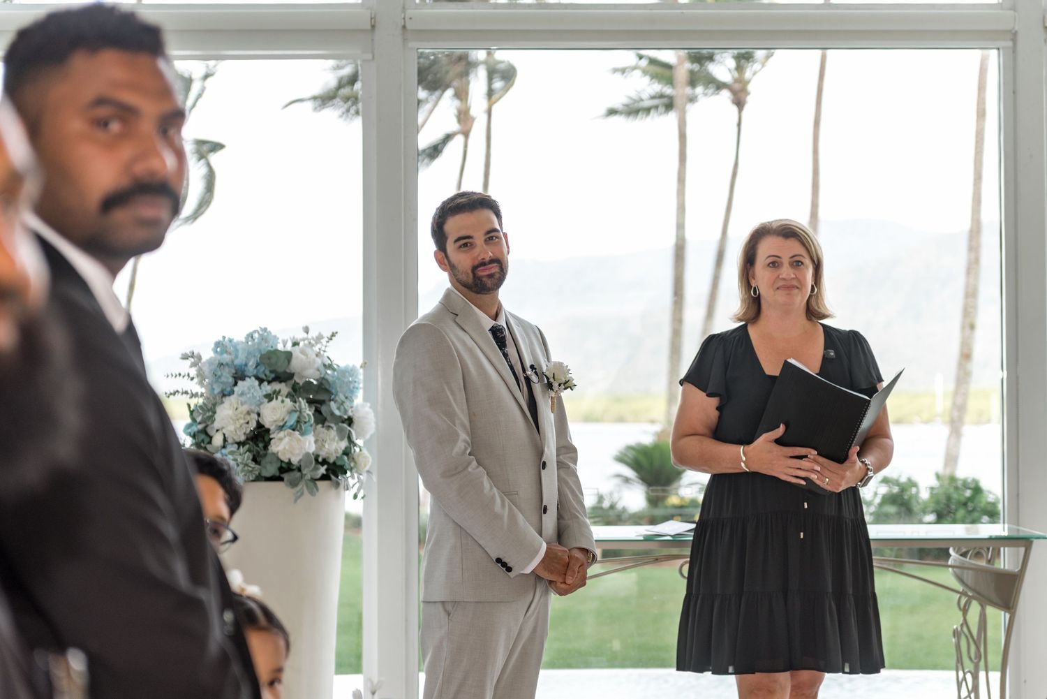 Groom in grey suit holds hands during wedding ceremony with celebrant reading vows.