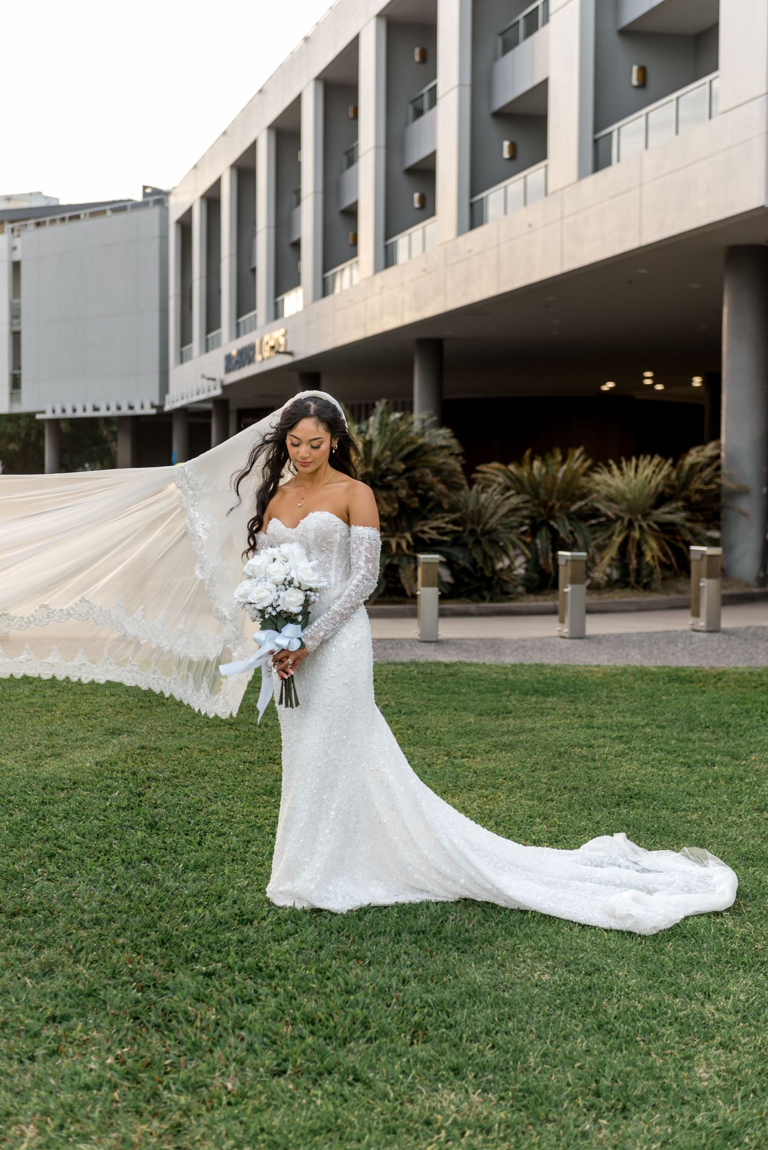 Bride in lace off-shoulder gown with flowing veil and white bouquet standing on green lawn outside modern building.