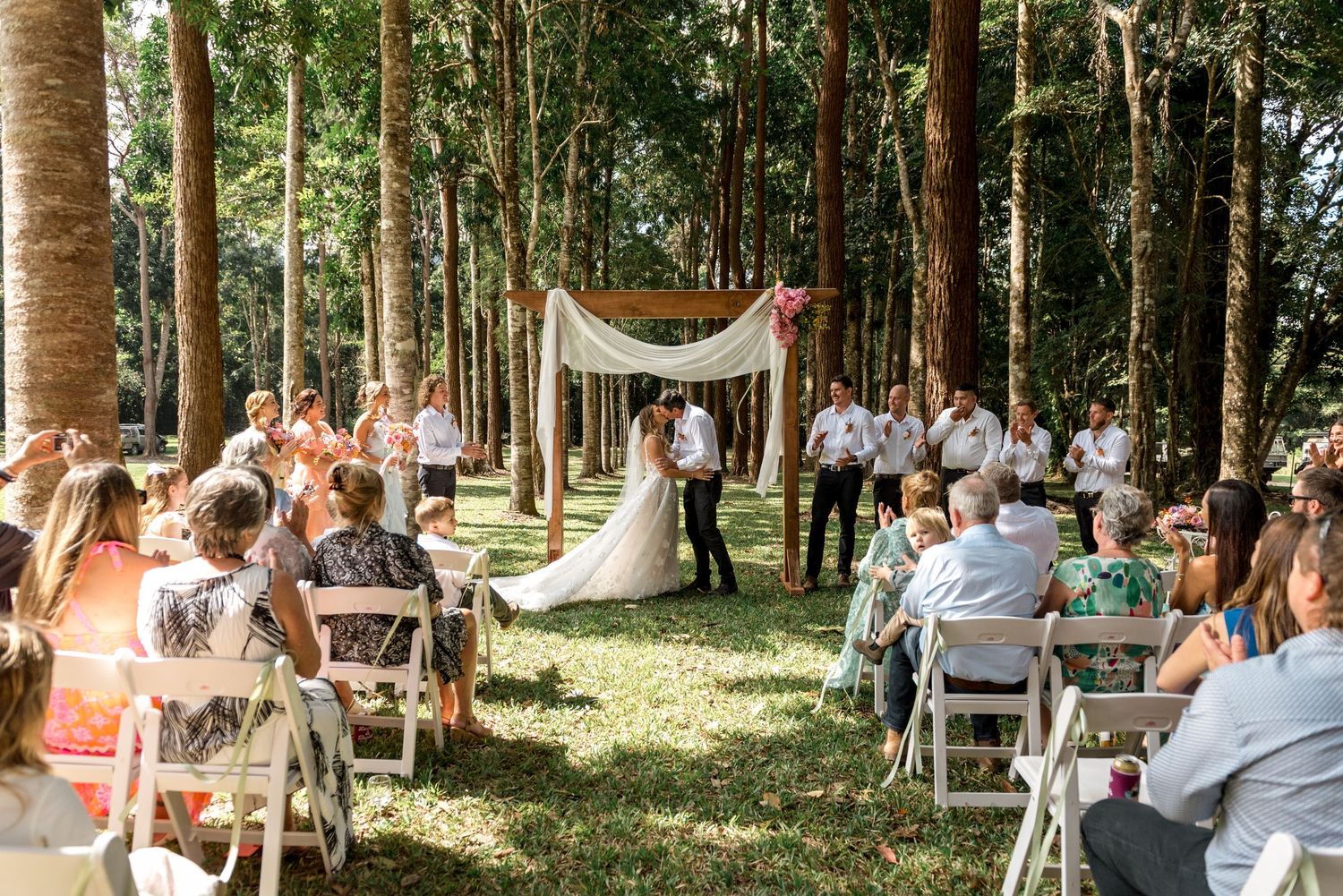 Outdoor wedding ceremony in a forest with guests seated as bride and groom kiss under a draped wooden arch.