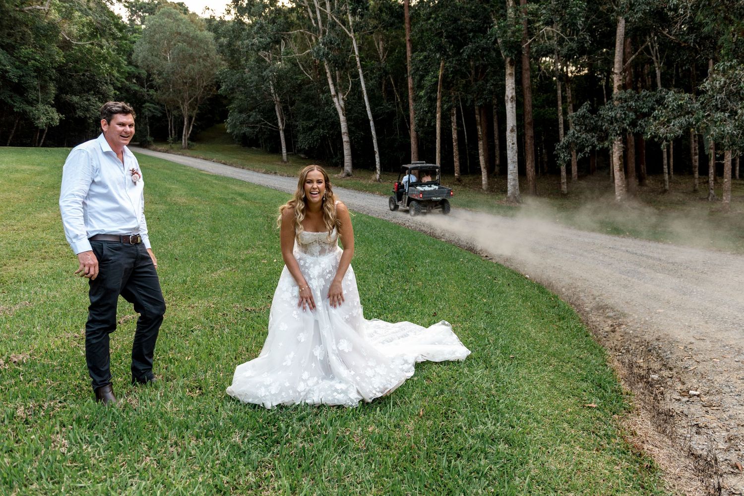 Bride and groom standing on green grass beside a dusty gravel road with a vehicle driving past tall trees.
