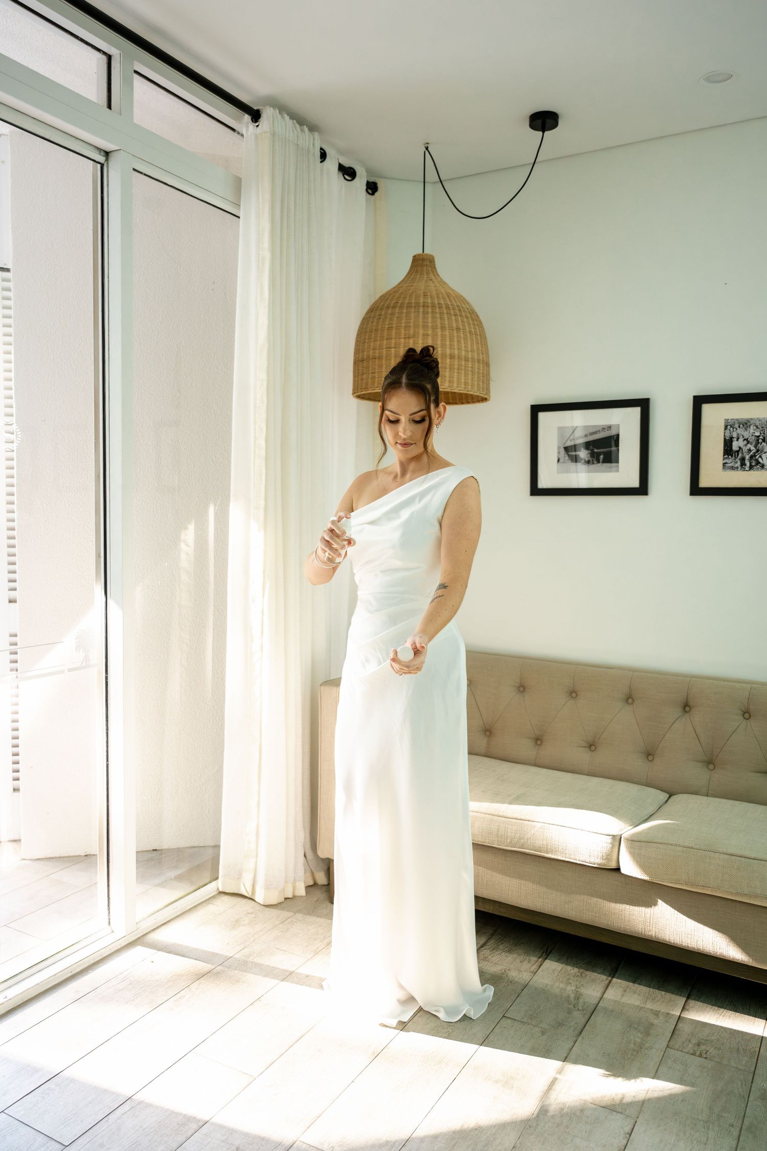 Bride in sleek one-shoulder white wedding gown stands by sunlit window in bright modern room.