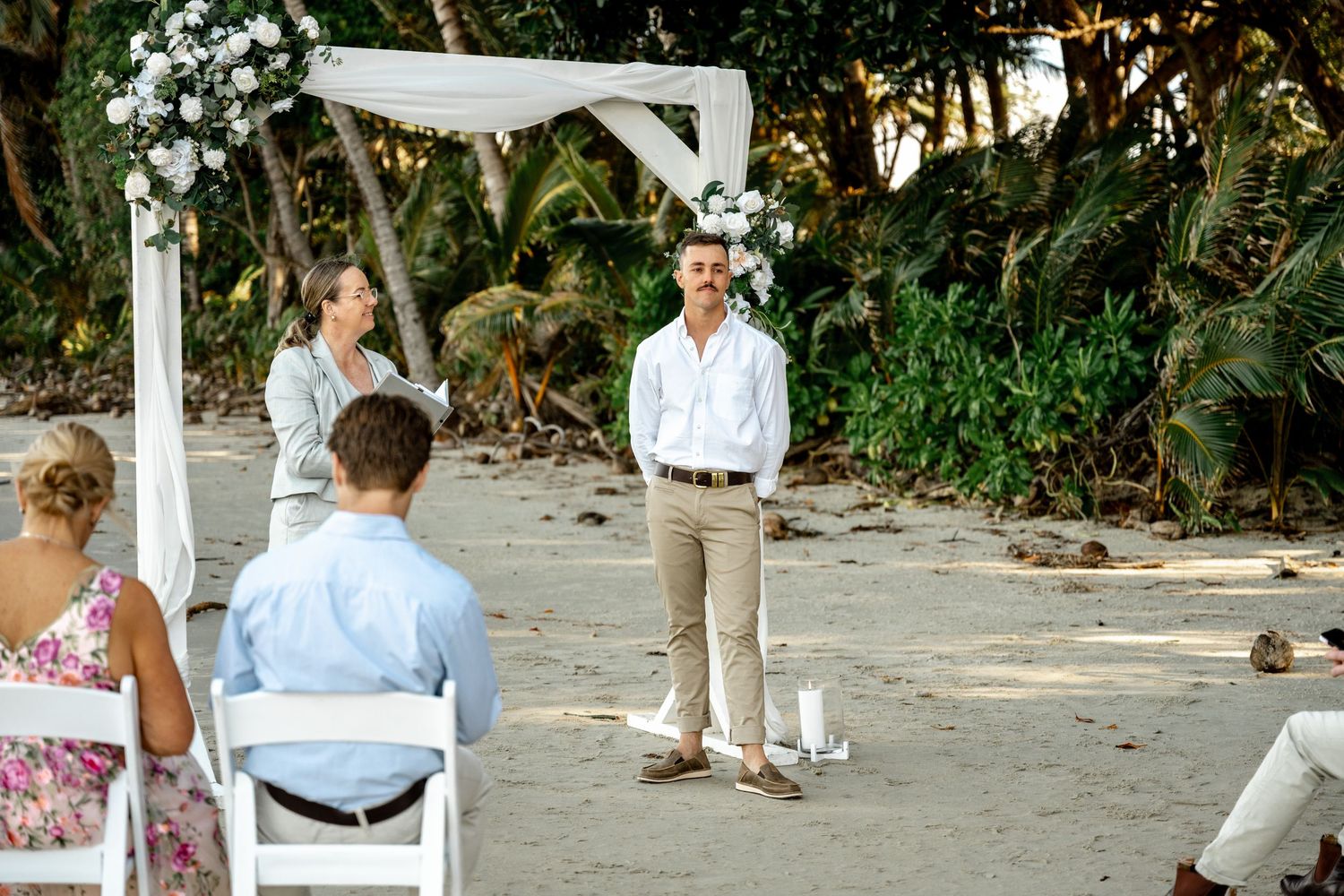 Groom standing at beach wedding altar decorated with white flowers and draping, guests seated on white chairs at Four Mile Beach.